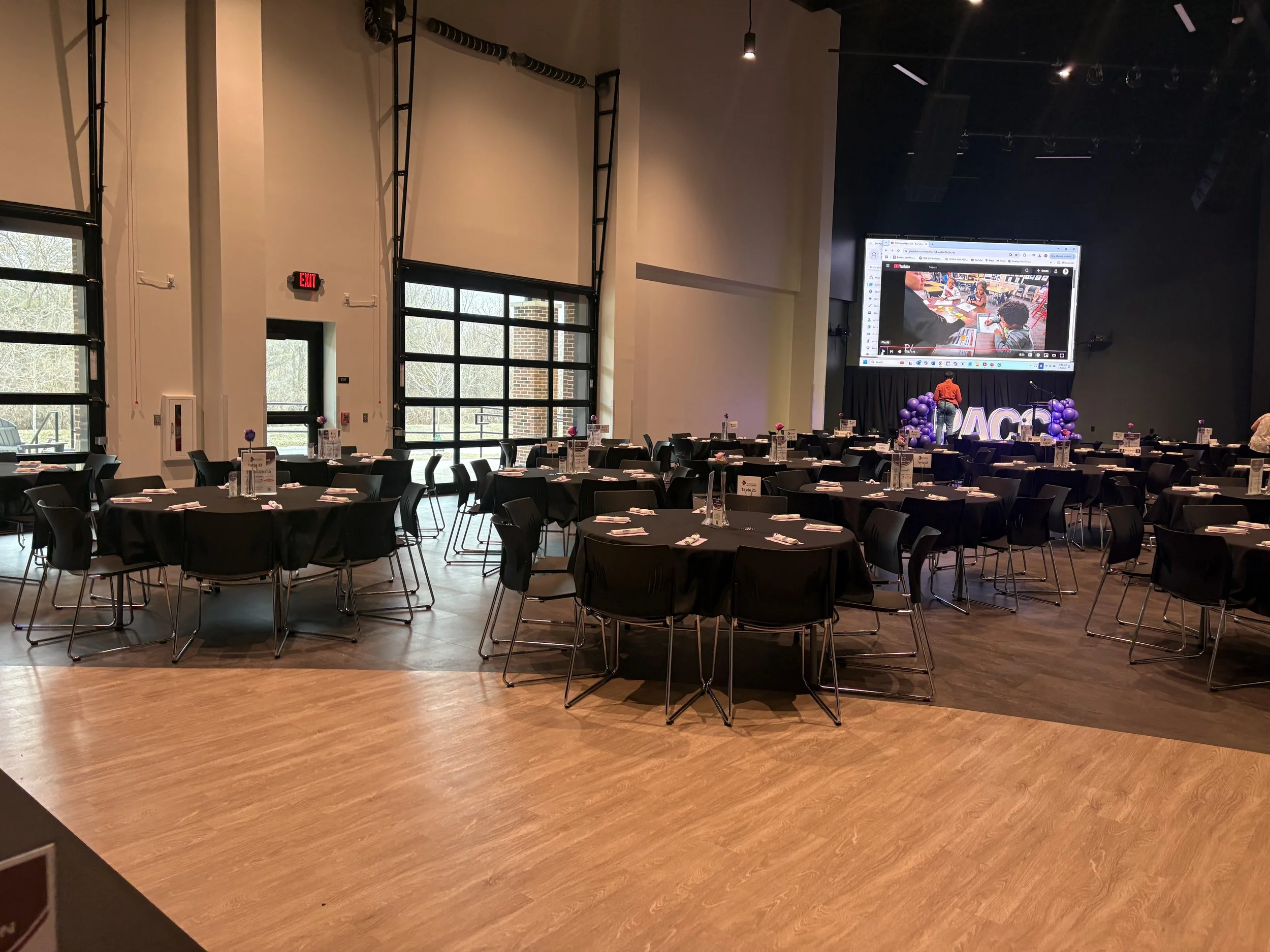 Event hall with round tables covered in black tablecloths, set with white napkins, and purple centerpieces. A stage with a large screen displays a video, with purple balloon decorations and illuminated letters spelling "PACE" at the front of the stag