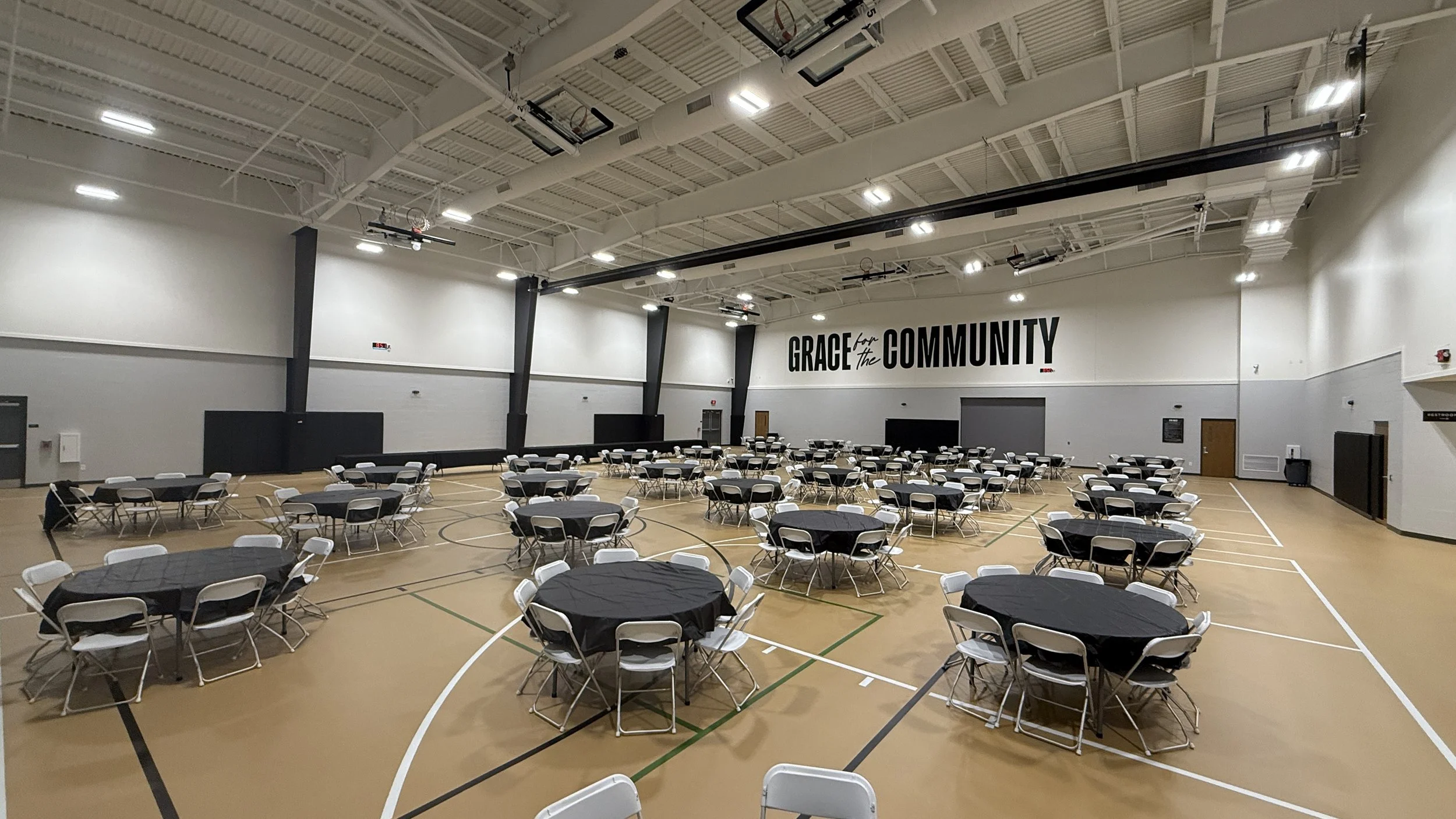 Large gymnasium with round tables and chairs set up for an event. The walls are white, and the ceiling is high with bright lights. A sign on the wall reads "Grace for the Community."