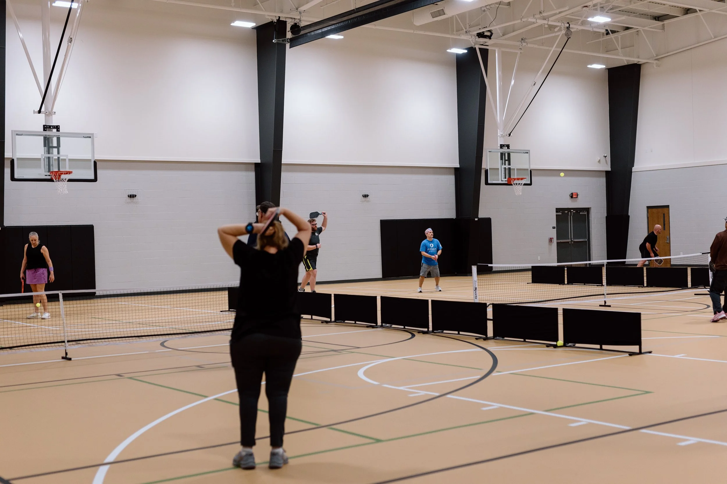 People playing pickleball on indoor courts with two basketball hoops visible on the walls.