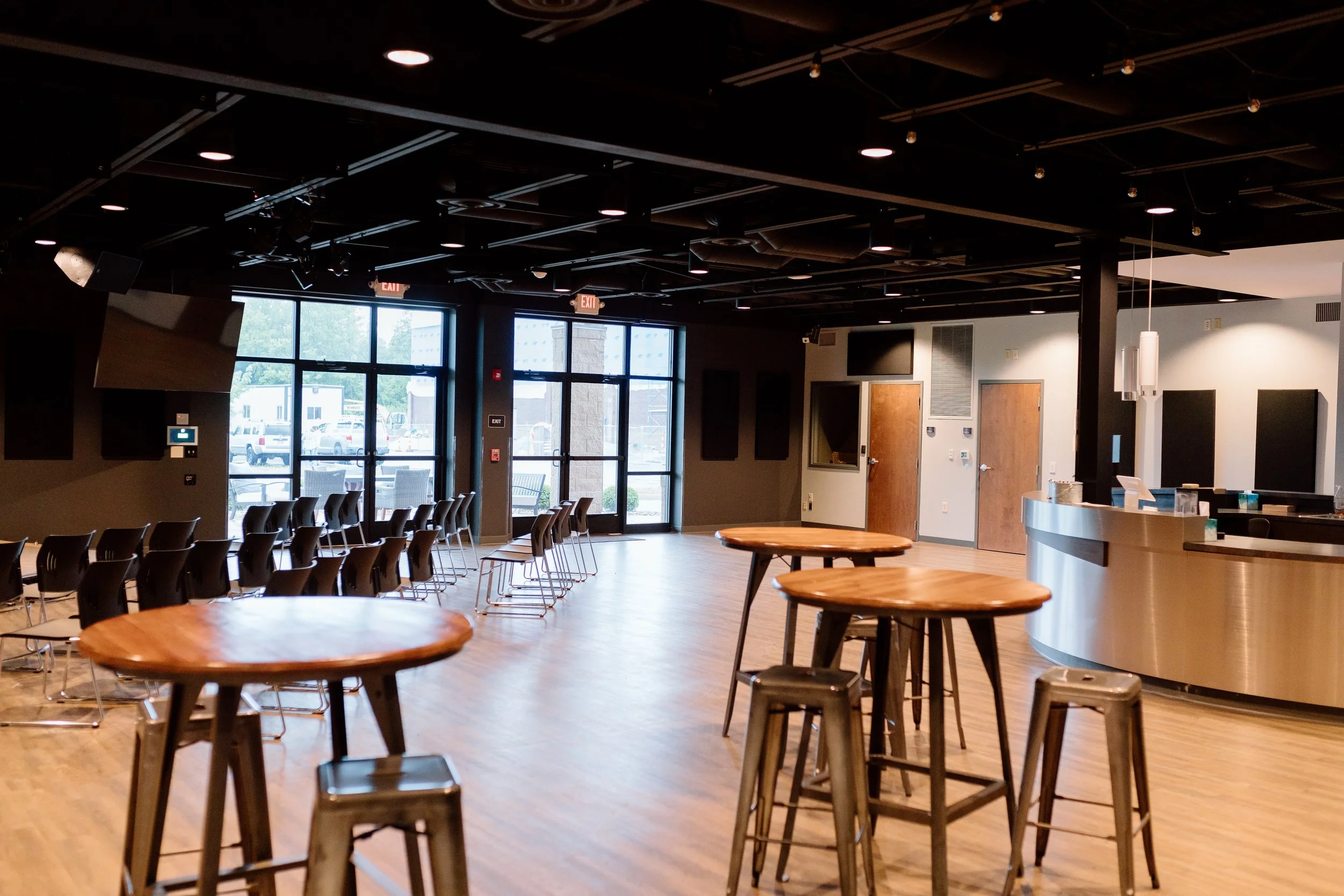 Interior of a modern cafe or lounge with high tables and stools, a curved service counter, chairs arranged near large glass windows, and a ceiling with spotlights and mounted speakers.