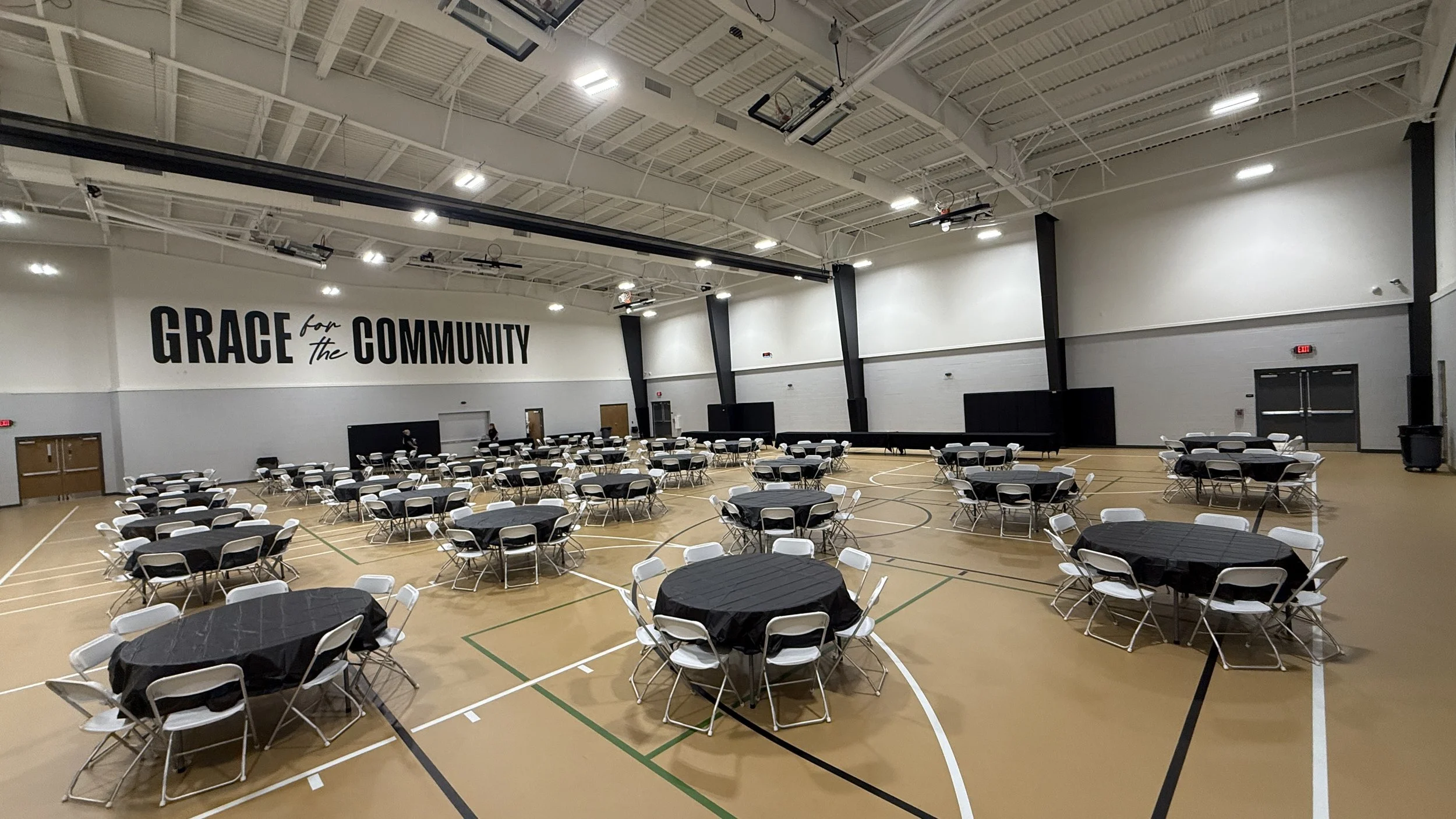 A large indoor gymnasium set up for an event with round tables covered in black tablecloths and white chairs. The walls feature a large sign that reads "GRACE for the COMMUNITY."