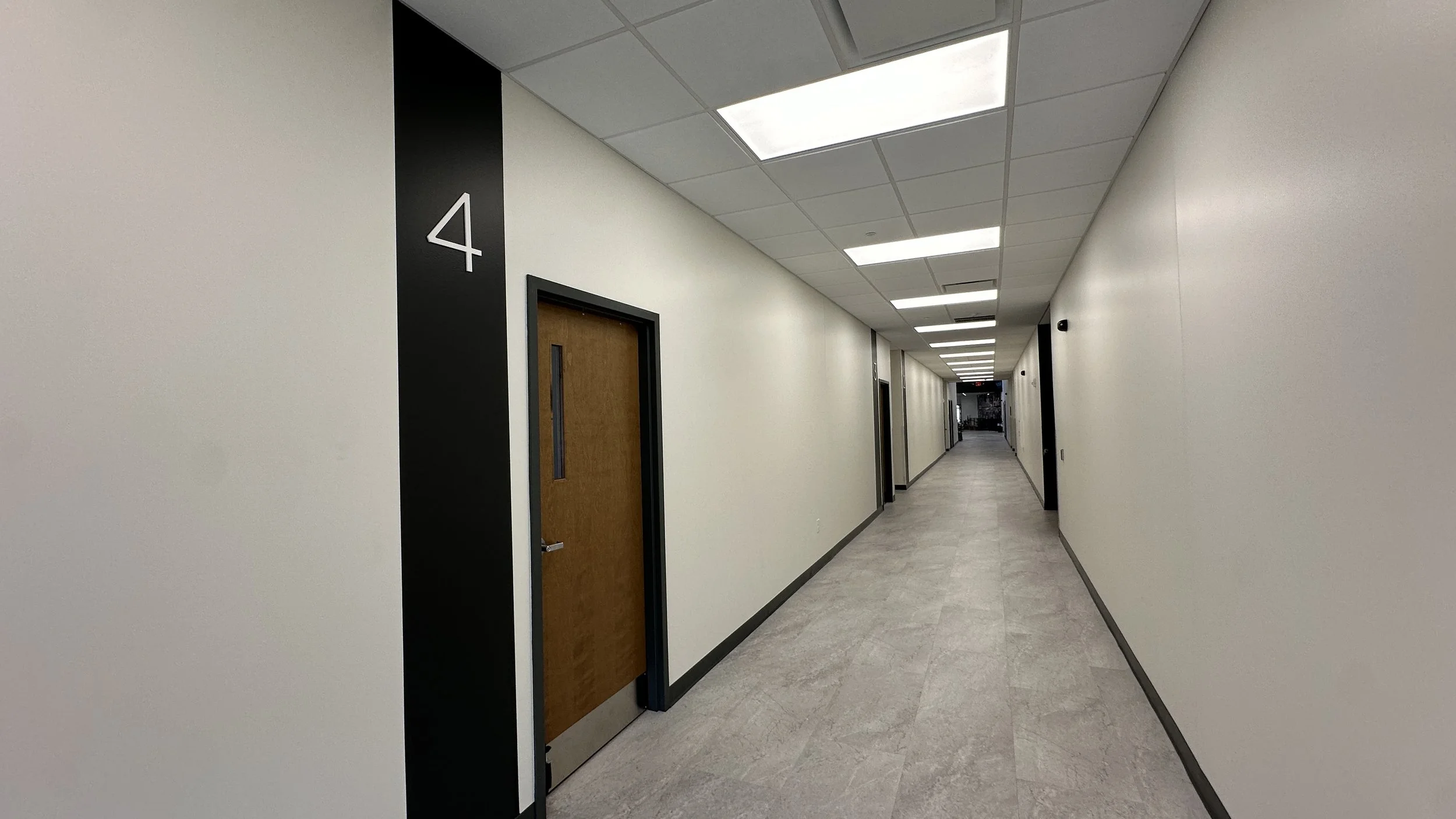 Empty beige-walled hallway in a building with a door labeled '4' on the left, fluorescent ceiling lights, and a view towards the end of the corridor.