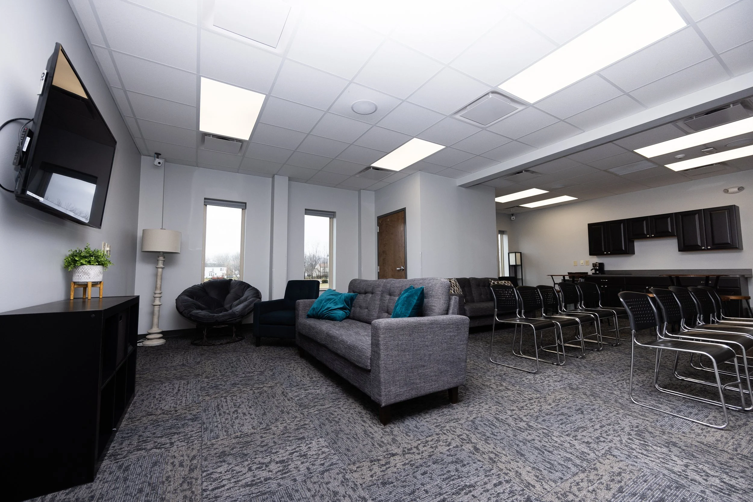 A spacious lounge with a gray couch, a black egg chair, a side table with a plant, a wall-mounted TV, and a kitchenette with black cabinets and multiple chairs.
