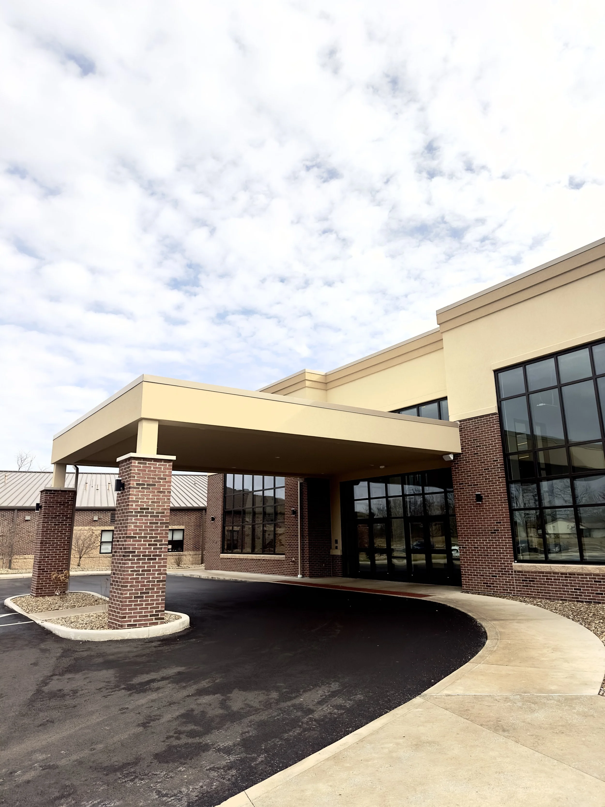 Driveway leading to the entrance of a modern brick building with large glass windows and a covered drop-off area.