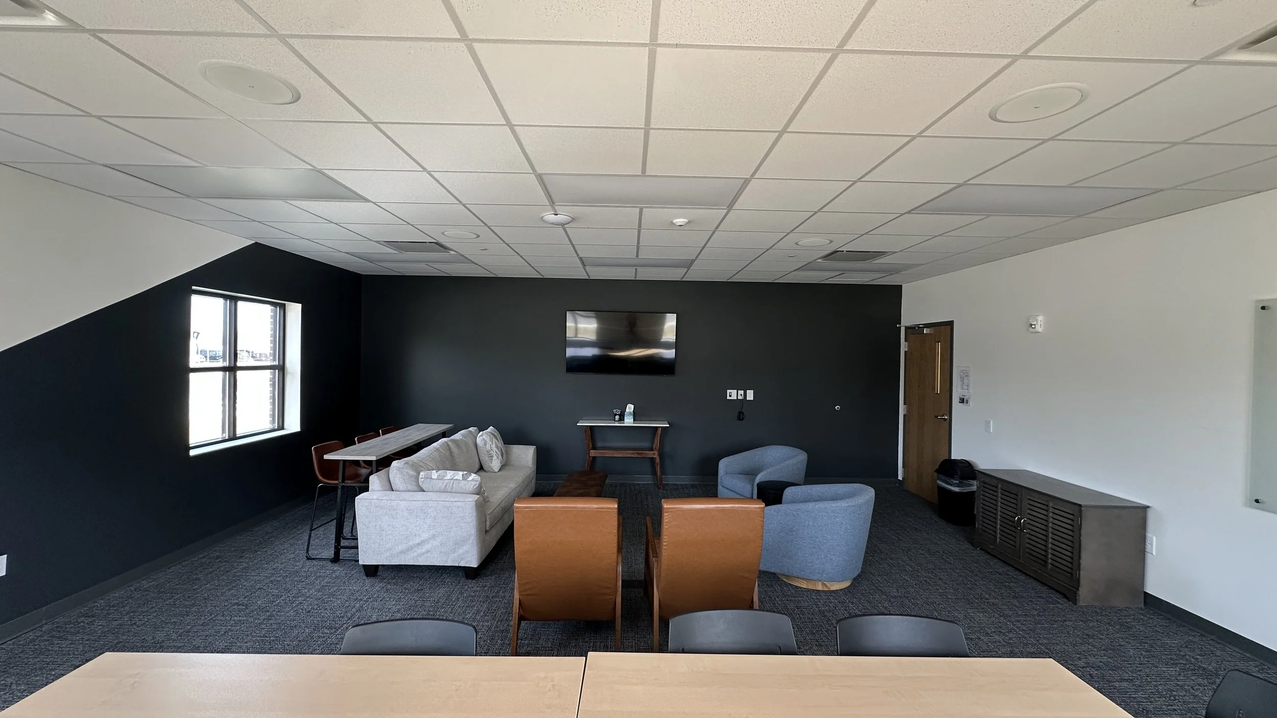 Empty lounge area with chairs, sofas, a wall-mounted TV, a window, a table, and a gray cabinet.