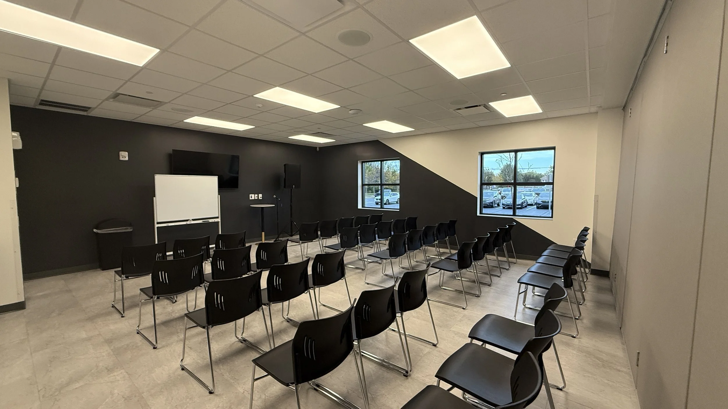 Empty conference room with black chairs arranged in rows facing a whiteboard and a television on a dark wall. Windows on the right side overlook a parking lot with cars.