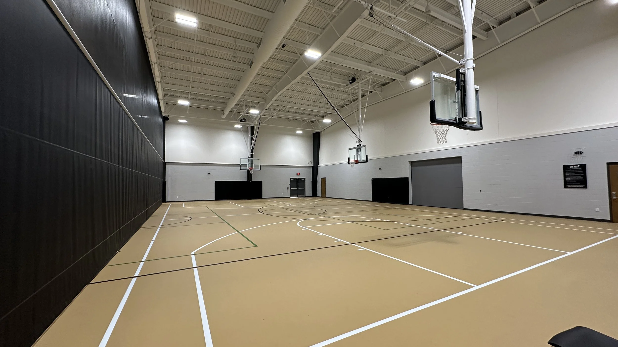 Interior view of a spacious indoor basketball court with multiple hoops, light-colored polished floor, high ceiling, and walls painted gray and black.