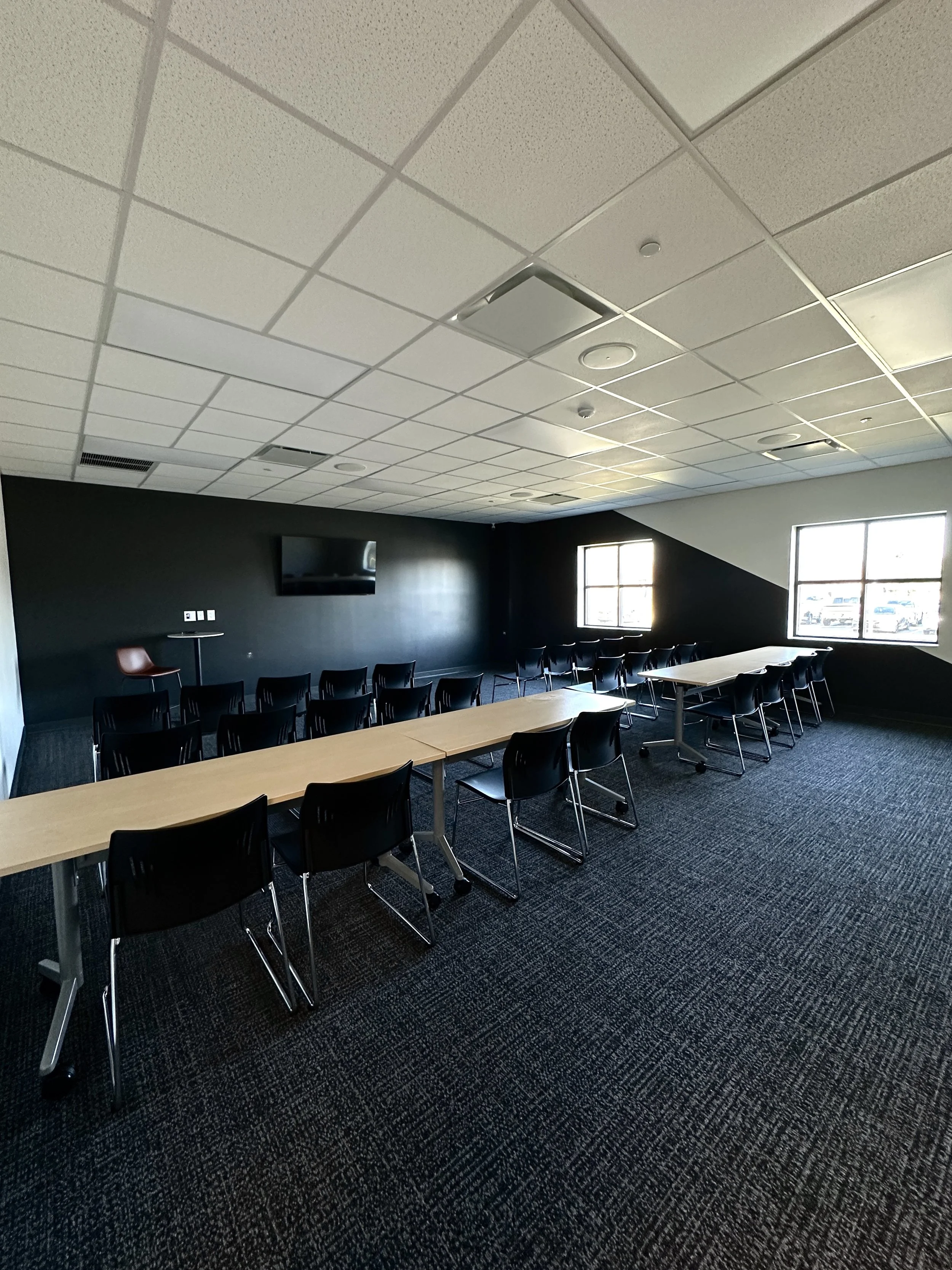 Empty conference room with two long tables, black chairs, a wall-mounted TV, and two windows letting in natural light.