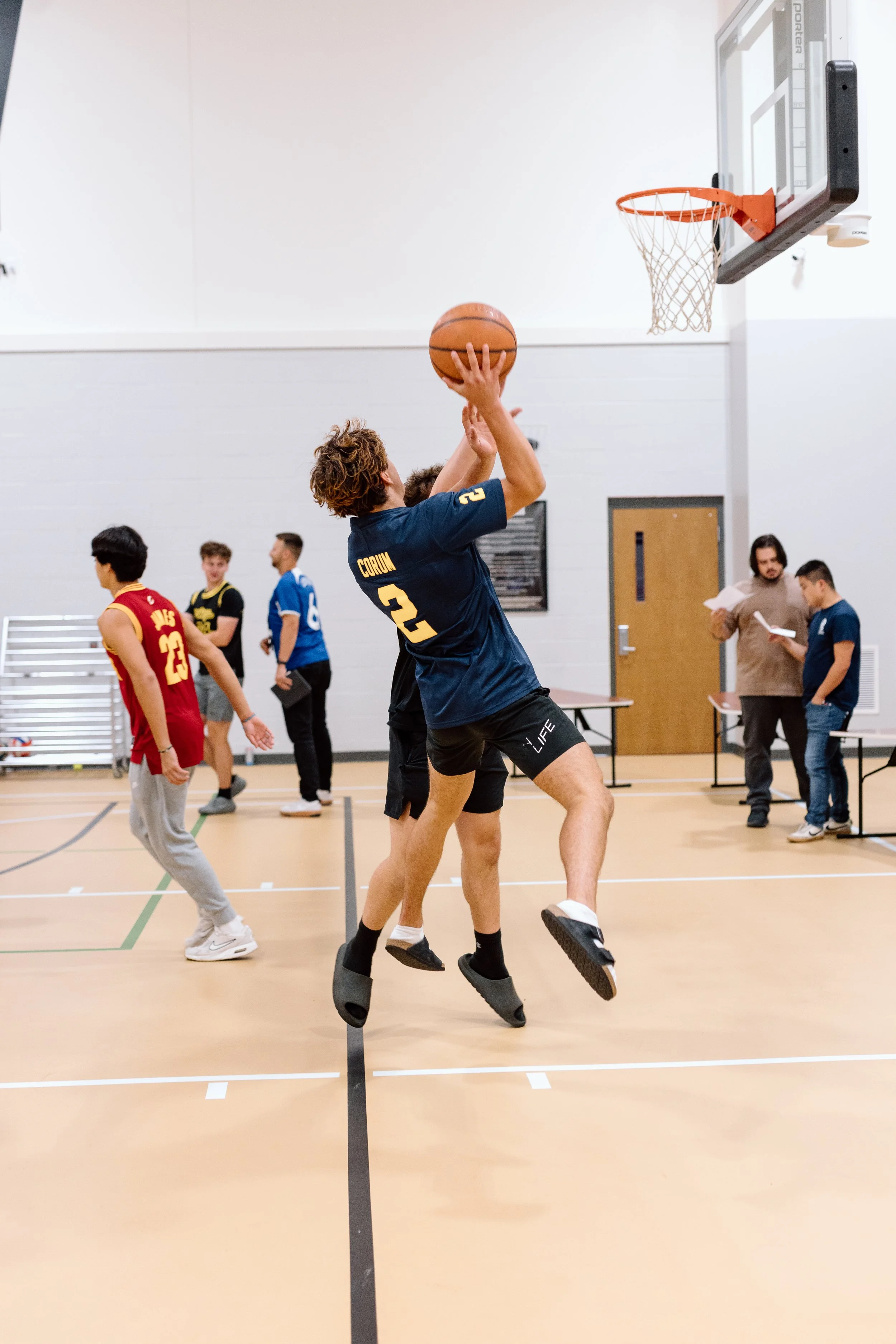 A person with pogo sticks is jumping and shooting a basketball towards the hoop during a basketball game or practice in an indoor gymnasium.