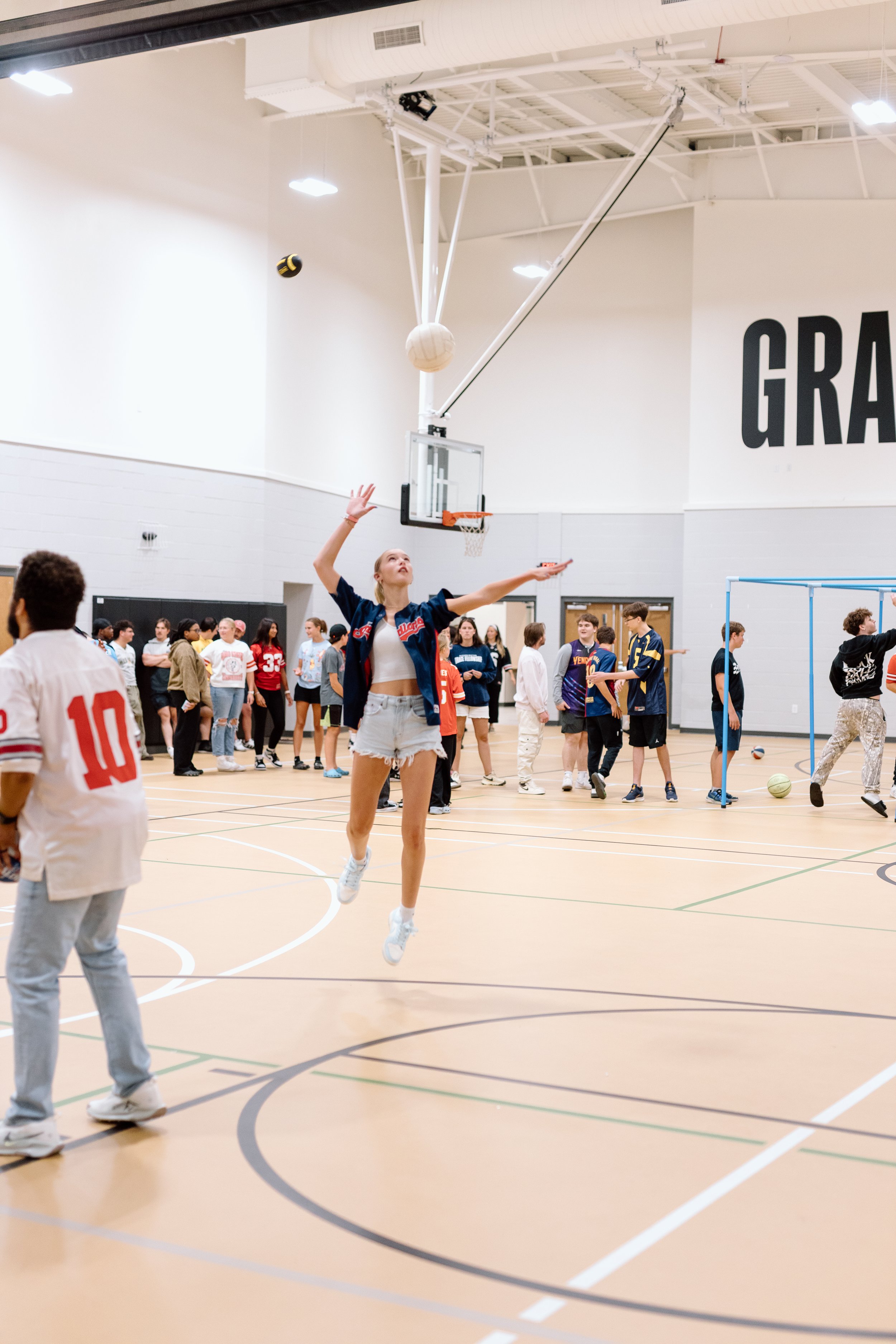 A girl in white shorts and a jacket jumping in an indoor gymnasium while playing basketball, with other kids and teenagers in the background.