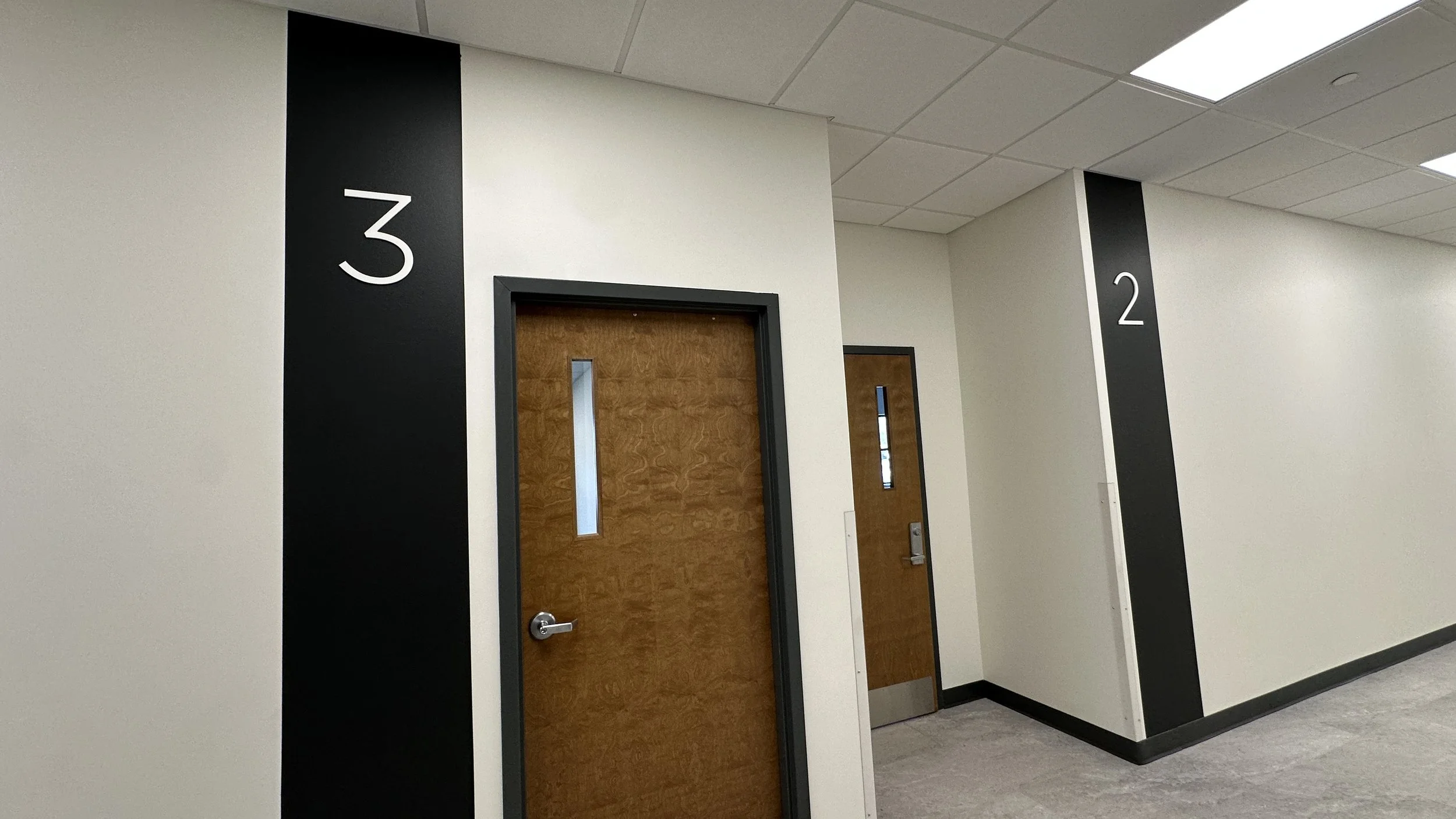 Hallway with two wooden doors numbered 2 and 3, white walls with black vertical number signs, ceiling with fluorescent lights.