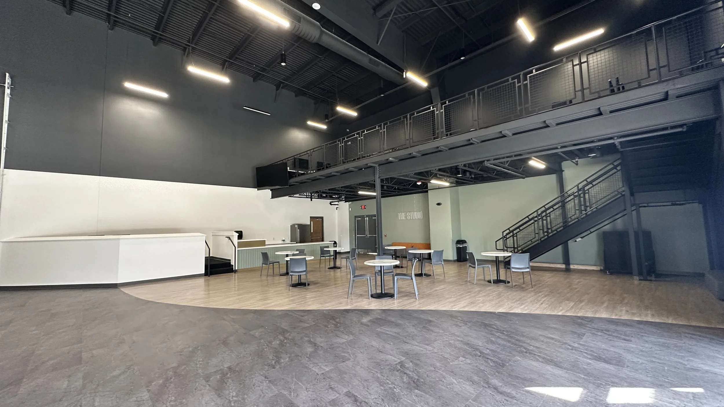 Empty indoor event space with high ceilings, black metal staircase, and several tables and chairs arranged for a gathering.