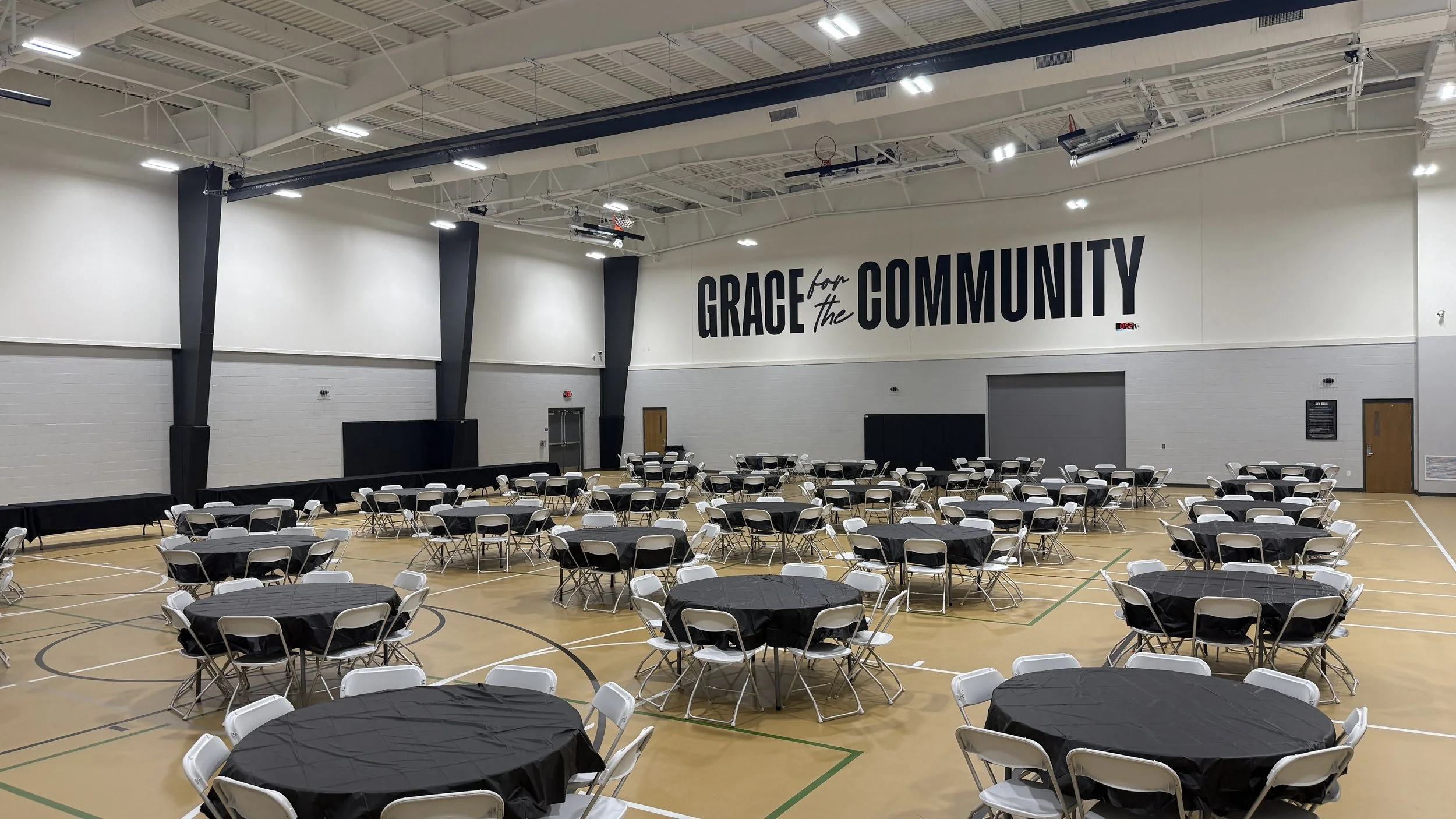 Large indoor gymnasium set up with round tables covered with black tablecloths and white folding chairs, with a large sign on the wall reading "Grace for the Community".