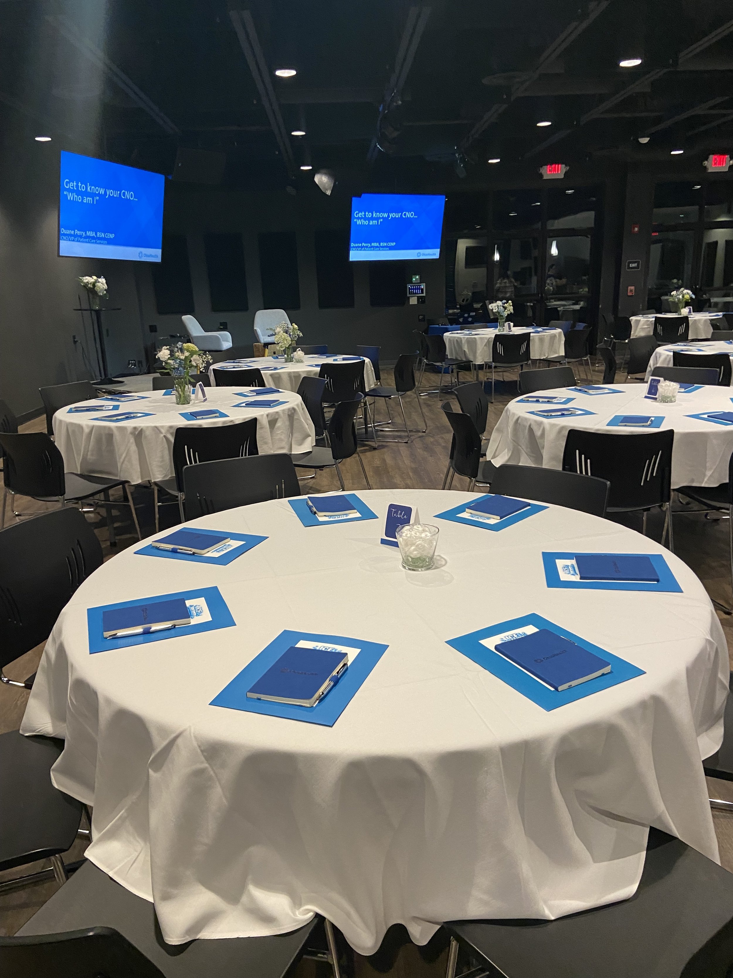 A conference room with round tables covered with white tablecloths. Each table has blue folders, pens, and a small centerpiece. The room has multiple chairs, two large screens displaying a presentation, and a step ladder in the background.