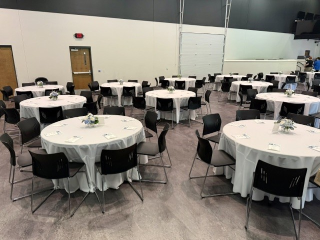 Multiple round banquet tables with white tablecloths and black chairs arranged in a large event space with minimal decoration.