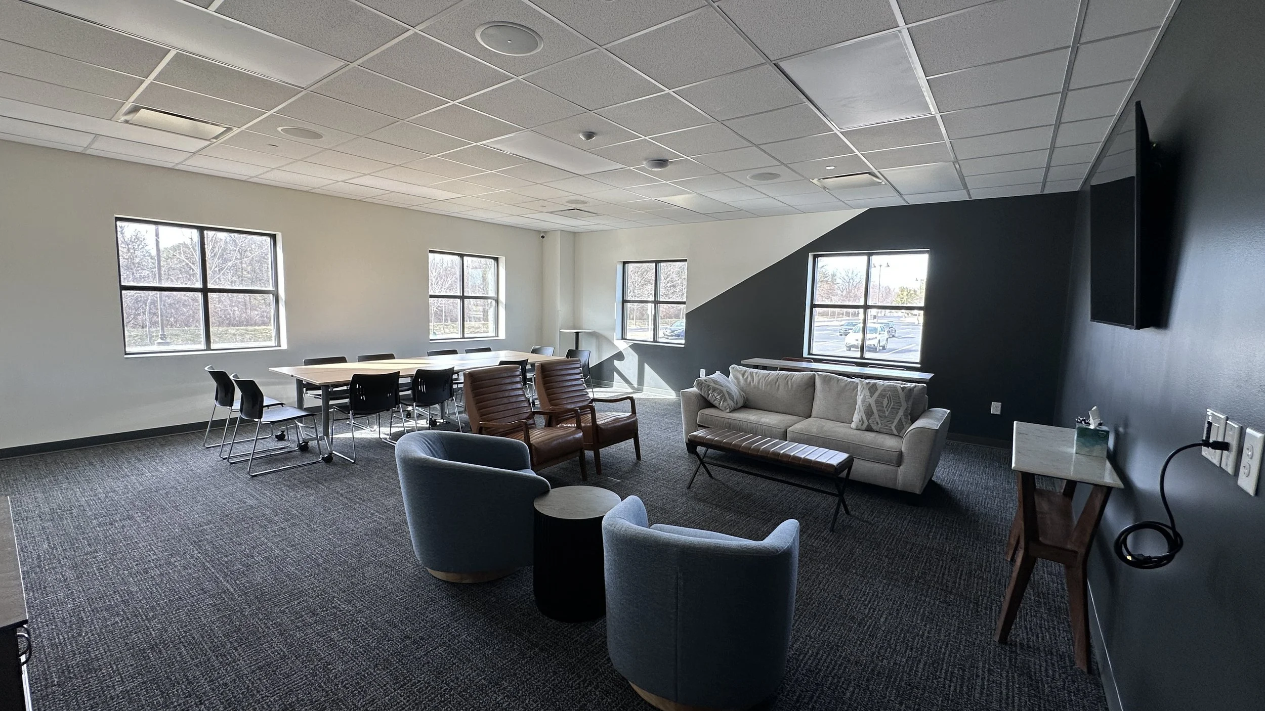 Interior of a modern lounge or meeting room with large windows, a long dining table with black chairs, a gray couch, two brown armchairs, blue armchair, small side tables, a TV on a dark wall, and a small table with a tissue box.