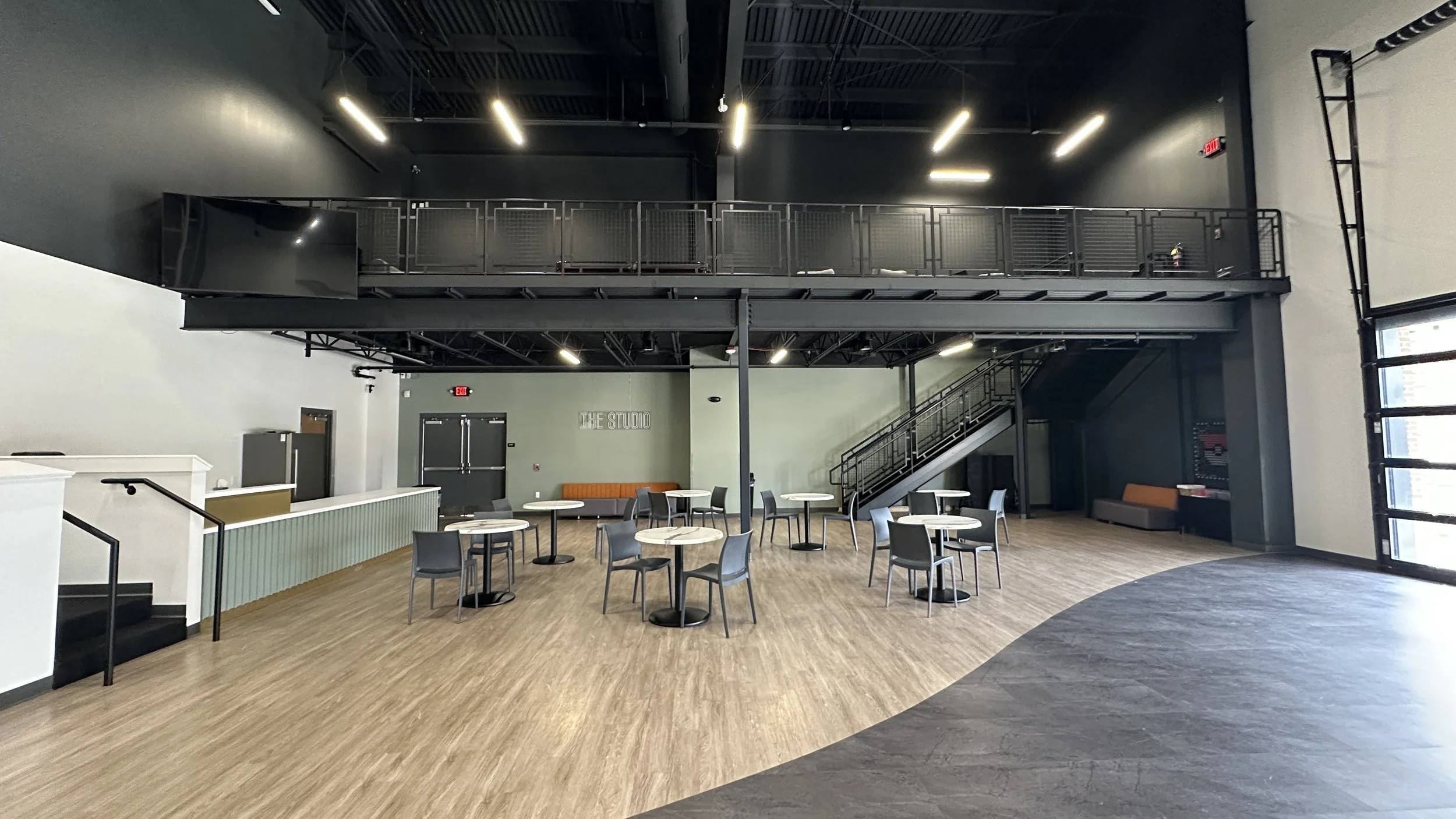 An empty modern studio lounge with round tables and chairs, a staircase to an upper level, and large windows letting in natural light.
