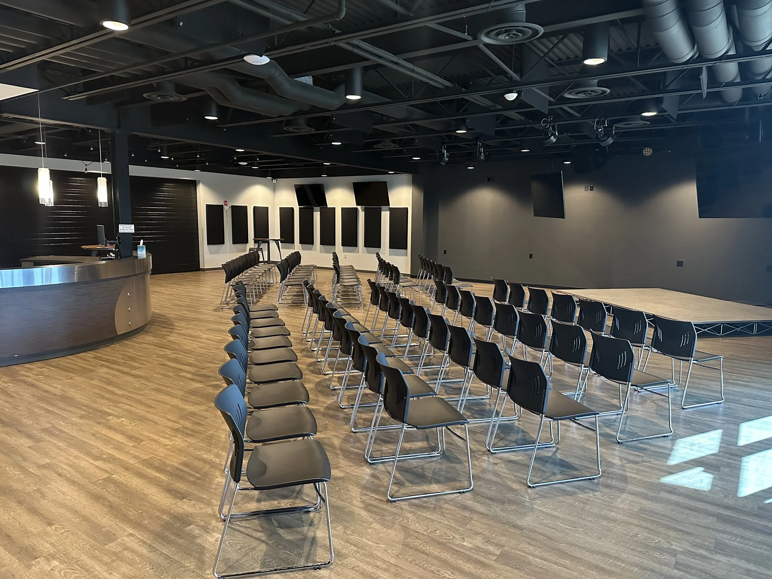 Empty conference room with black chairs arranged in rows, a small stage, and black acoustic panels on the walls, with wood flooring and ceiling with exposed ductwork.