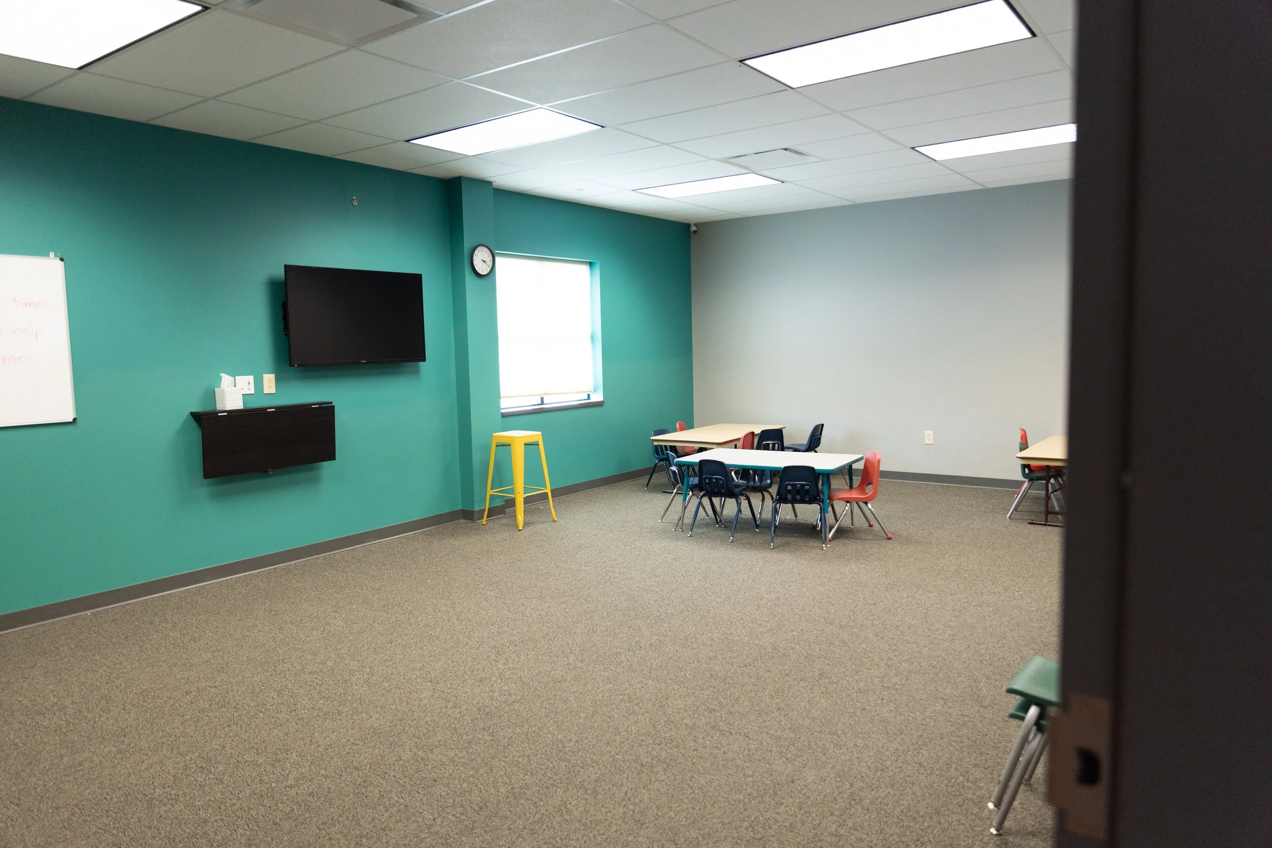 Empty classroom with tables and chairs, a large TV on the wall, a whiteboard, a window, and a wall clock.