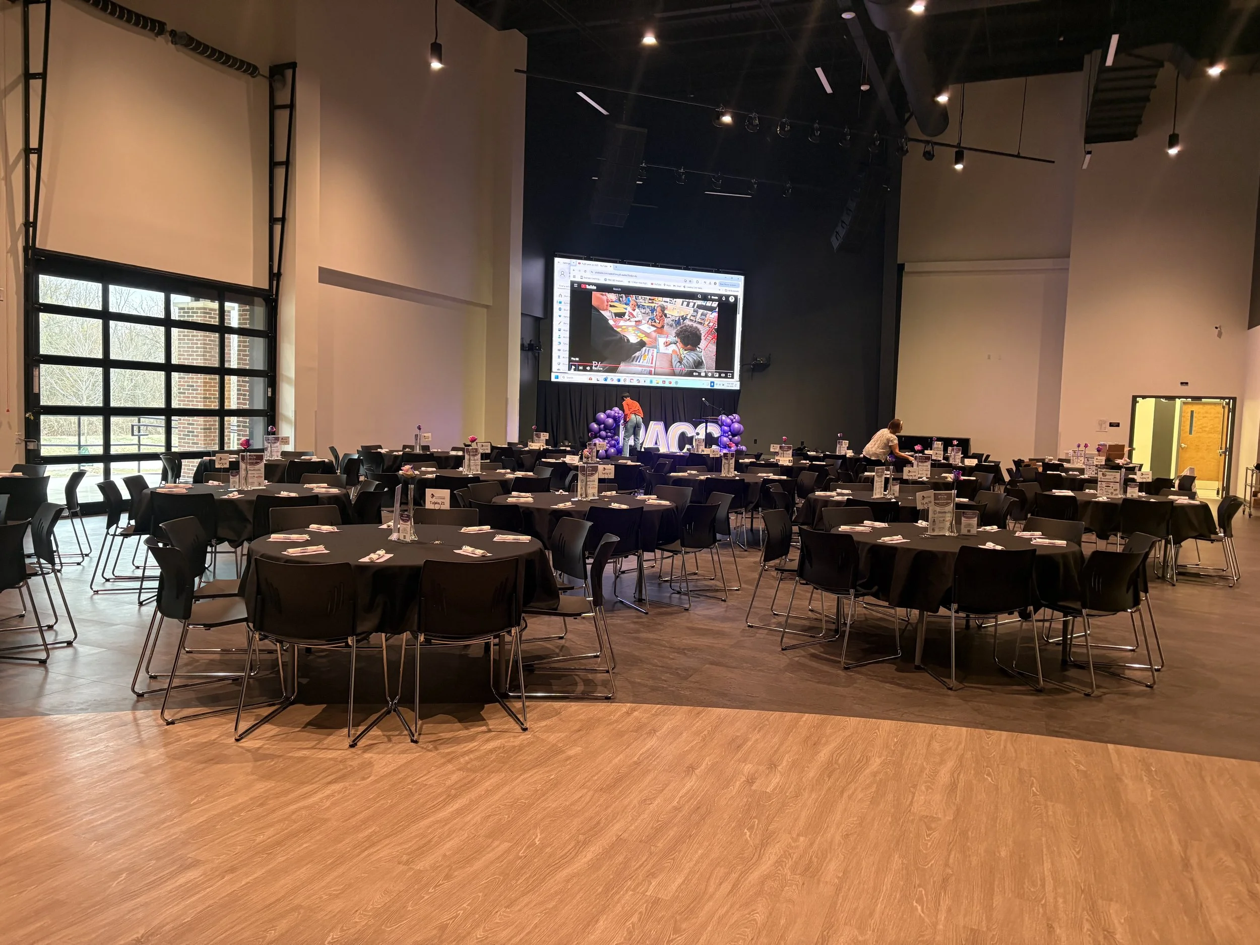 Event hall set up with round tables covered with black tablecloths, each with plates, napkins, and water bottles, arranged for a gathering or celebration, with a large stage at the front featuring decorated letters and balloons, a large screen displa