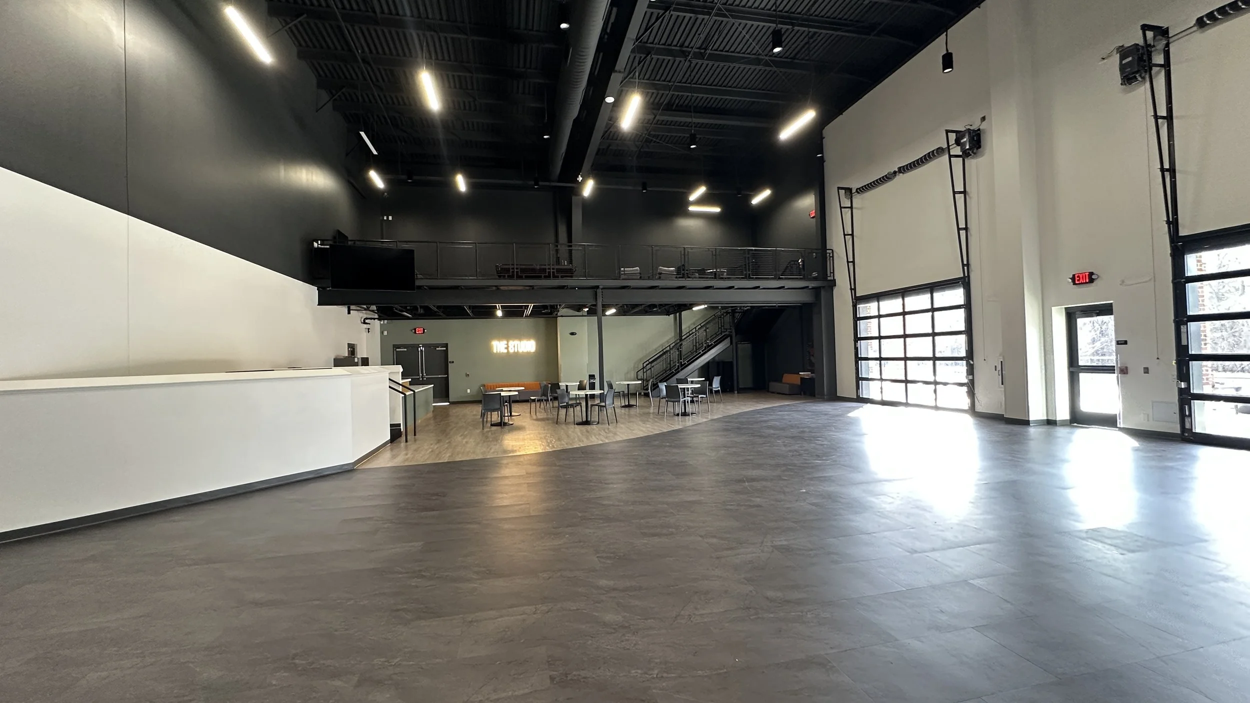 Spacious, modern event space with high ceilings, large windows, and minimal furniture. There are a few tables and chairs near the back wall, a staircase leading to an upper level, and a large garage-style door on the right.