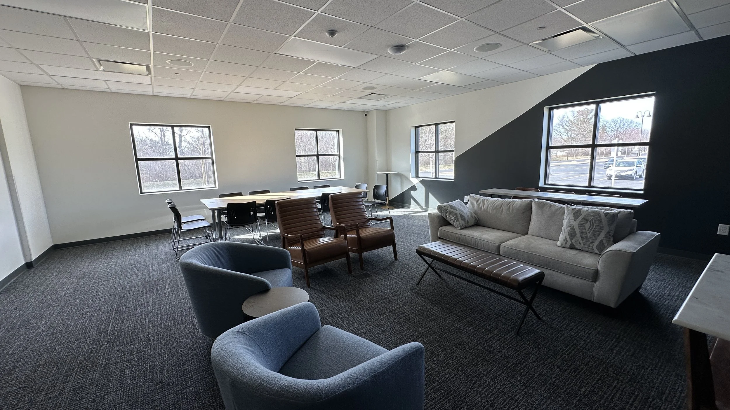 A spacious room with a gray carpet, white walls, and a black accent wall. It has large windows allowing natural light, a gray sofa with decorative pillows, two brown leather chairs, a blue armchair with an adjacent small round table, and a long dinin