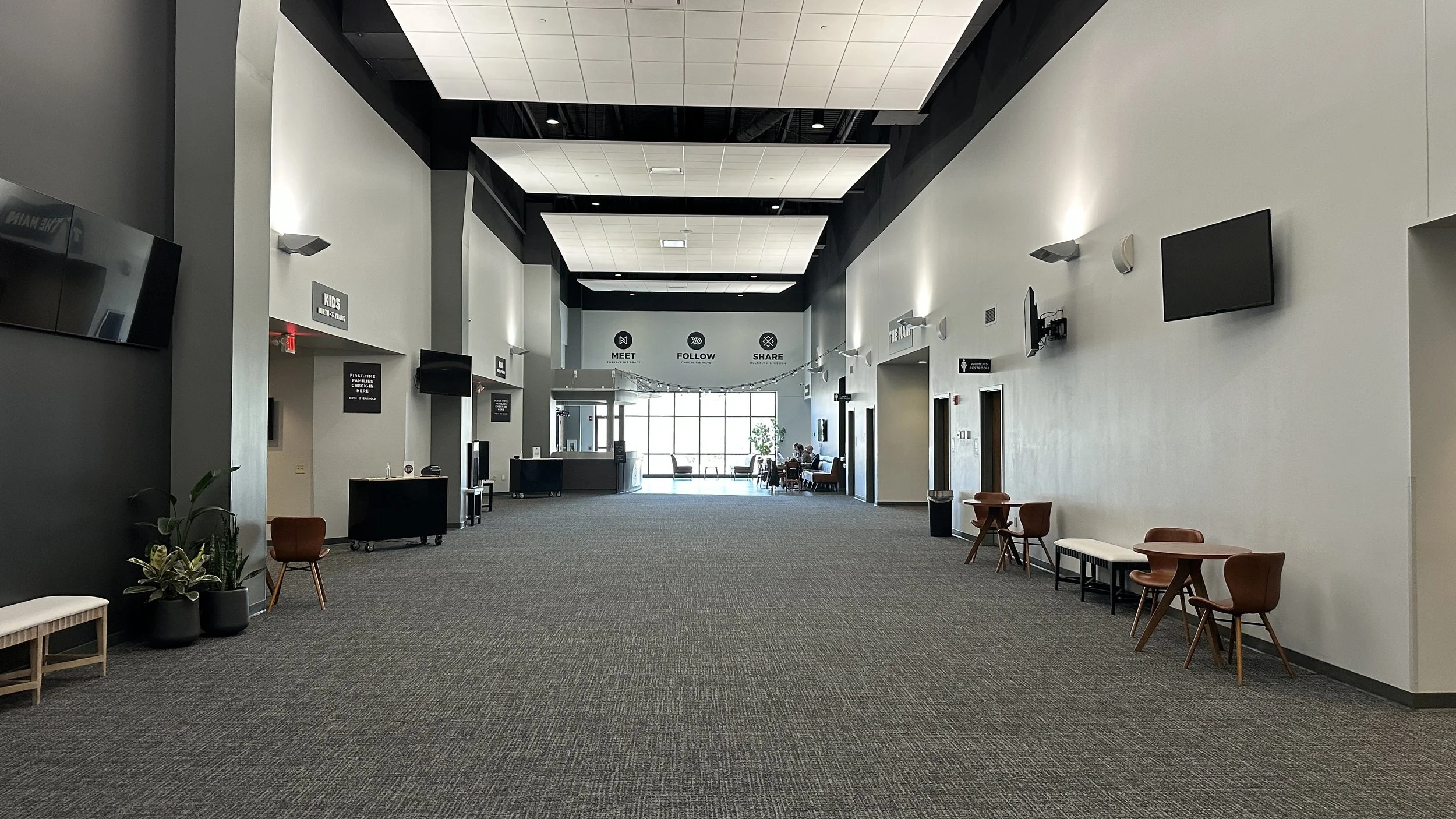 A spacious modern lobby with high ceilings, white and gray walls, and a gray carpeted floor. There are some chairs and small tables along the walls, a reception desk at the far end, and large windows allowing natural light to fill the space. Wall-mou