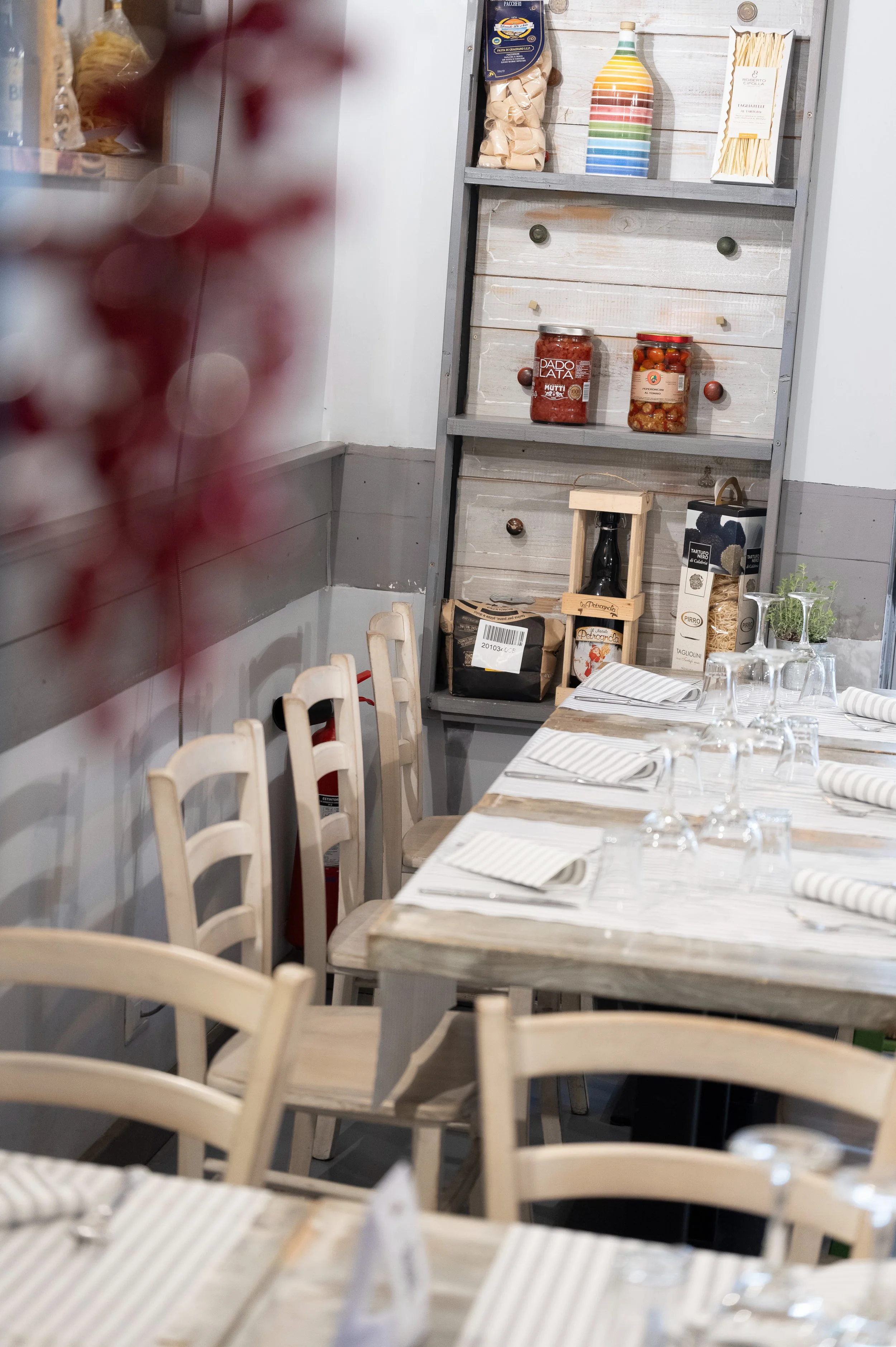 Interno di un ristorante con tavoli apparecchiati, sedie di legno, e una mensola con vari prodotti alimentari e oggetti decorativi sullo sfondo.