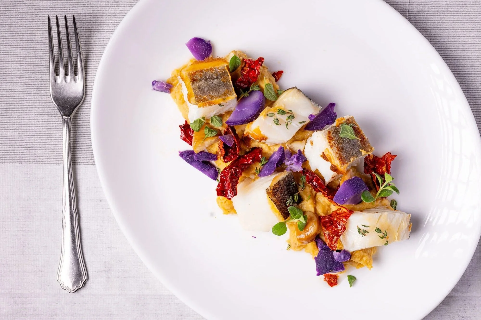 Piatto di baccalà, accompagnato da verdure e decorazioni di erbe aromatiche, servito su un piatto bianco con una forchetta di fianco.