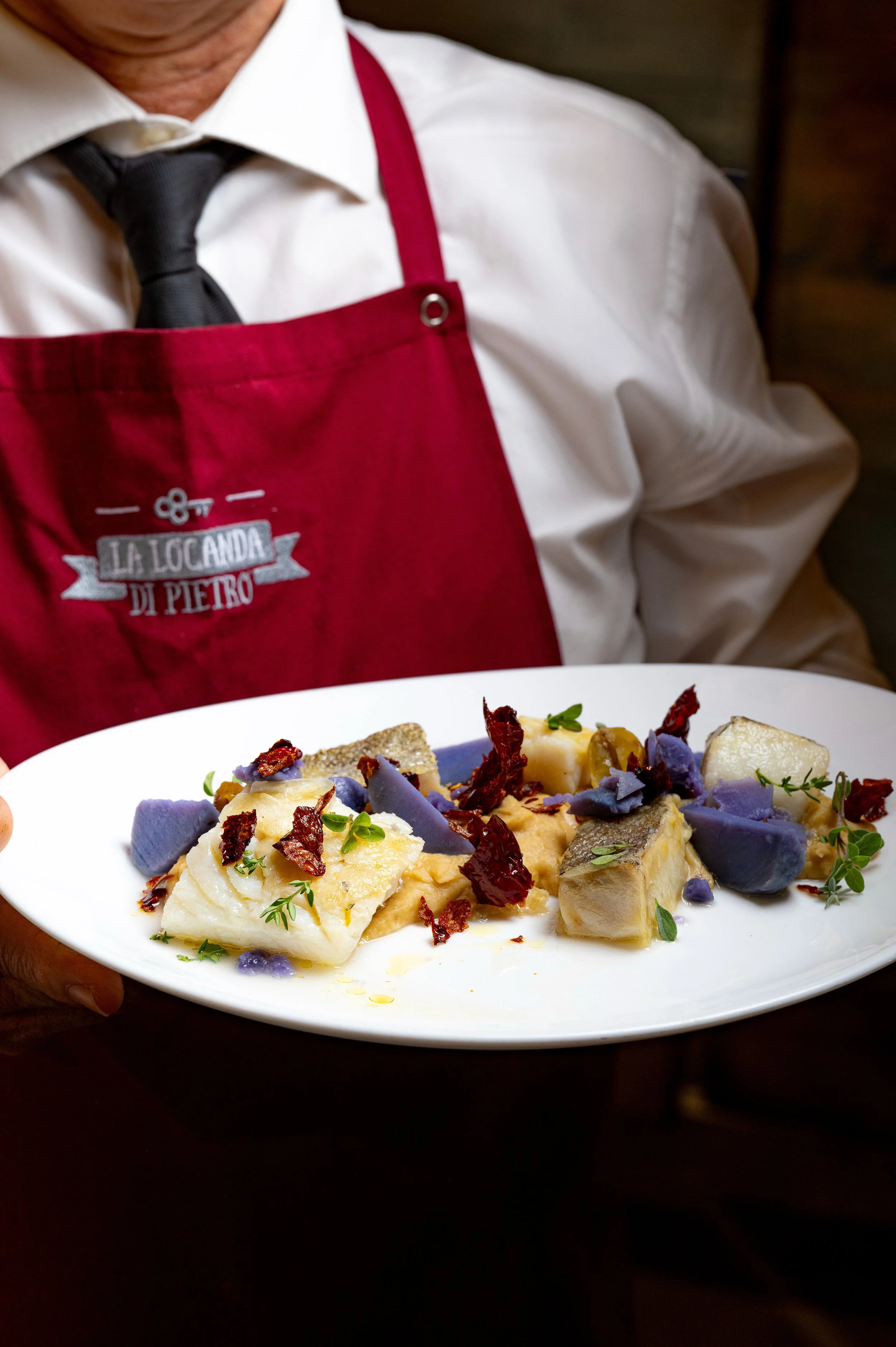 Cameriere che indossa una camicia bianca e un grembiule bordeaux che tiene un piatto con un piatto di pesce decorato con erbe, peperoni secchi e verdure colorate.