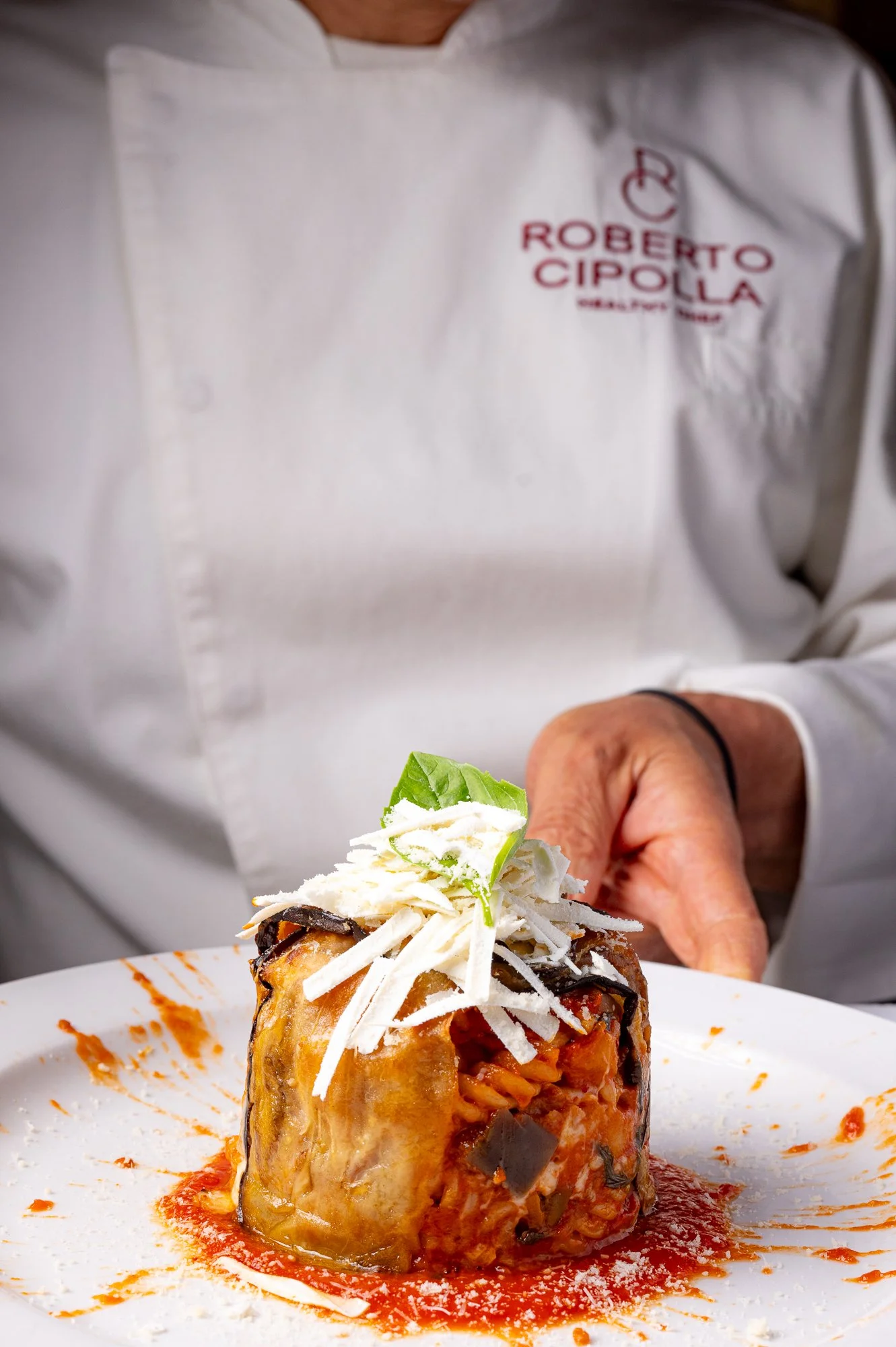Uno chef in giacca bianca con il logo 'Roberto Cipolla' sta servendo un piatto di timballo di melanzane, decorato con ricotta salata, salsa di pomodoro e un rametto di basilico.