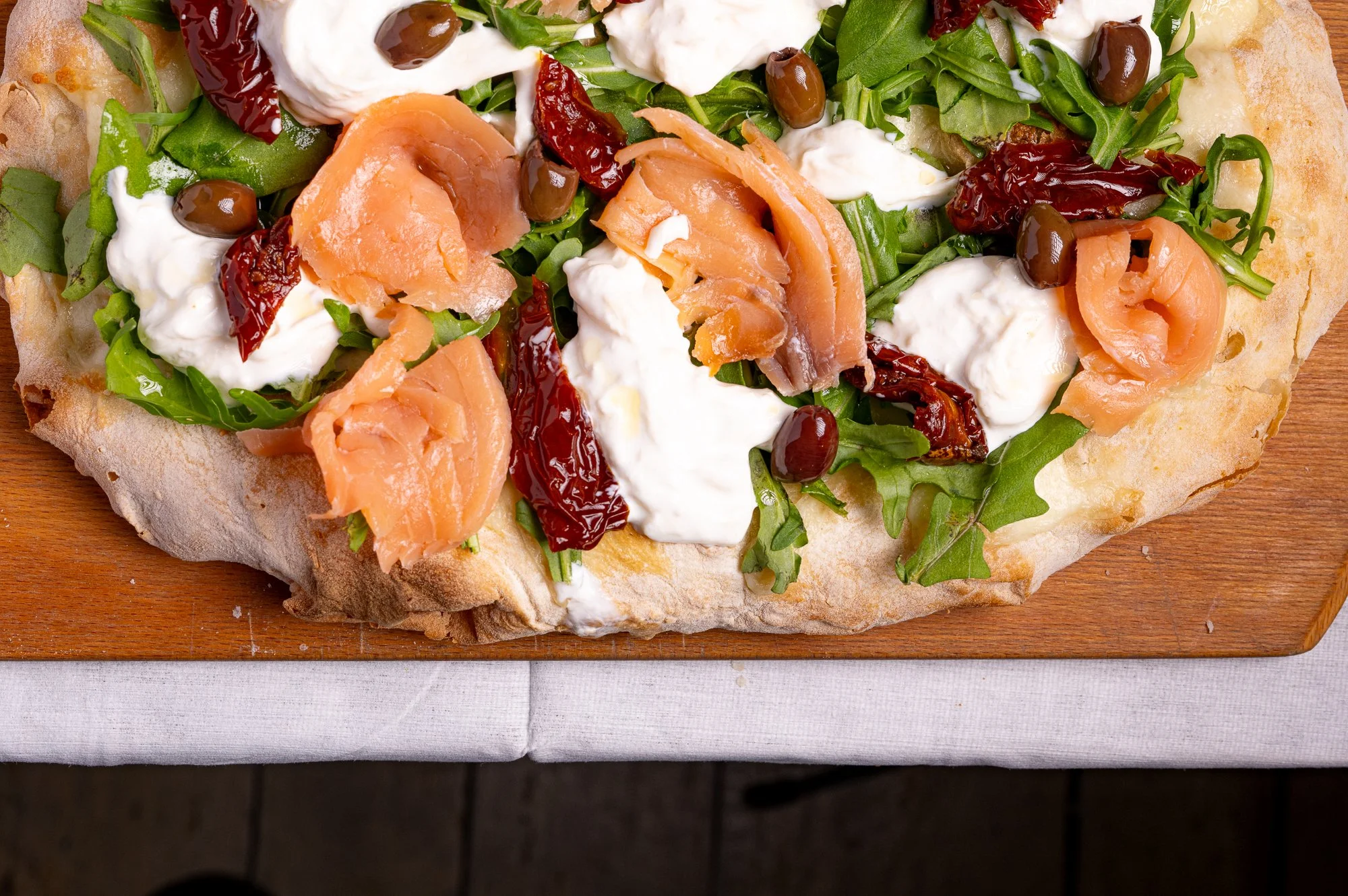 Fetta di pizza con salmone affumicato, pomodori secchi, stracciatella, rucola e olive nere su un tagliere di legno.