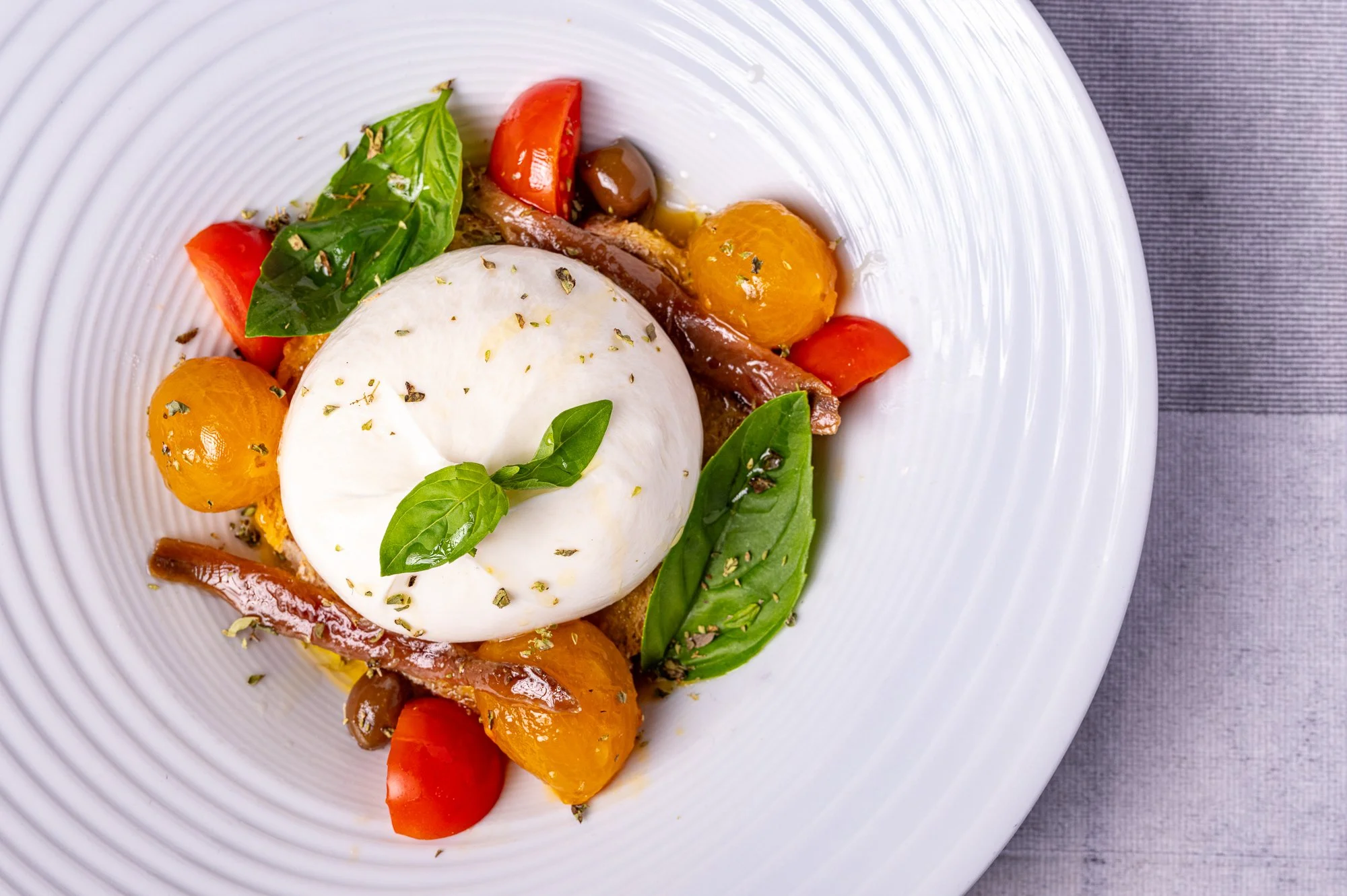 Piatto con burrata, pomodori, acciughe, foglie di basilico, olive e erbe aromatiche.