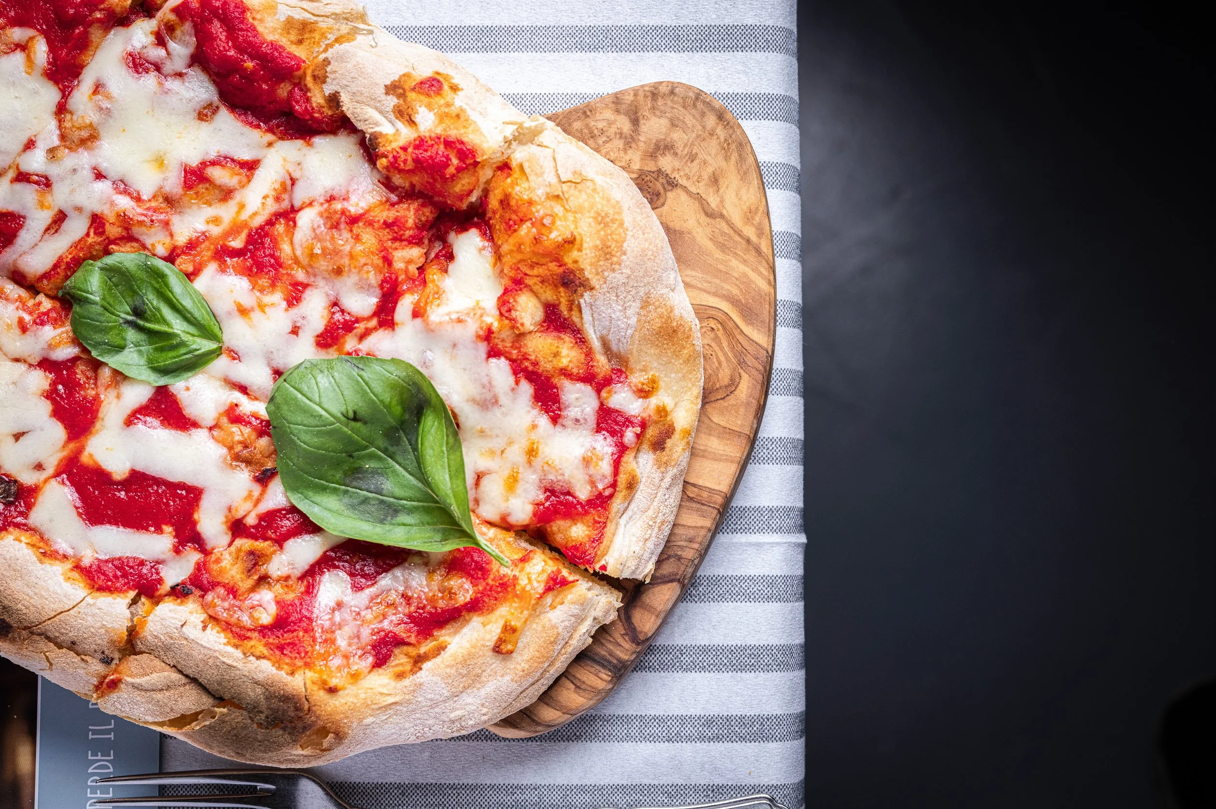 Pizza margherita con basilico fresco su un tagliere di legno, vista dall'alto su una tovaglia a righe.