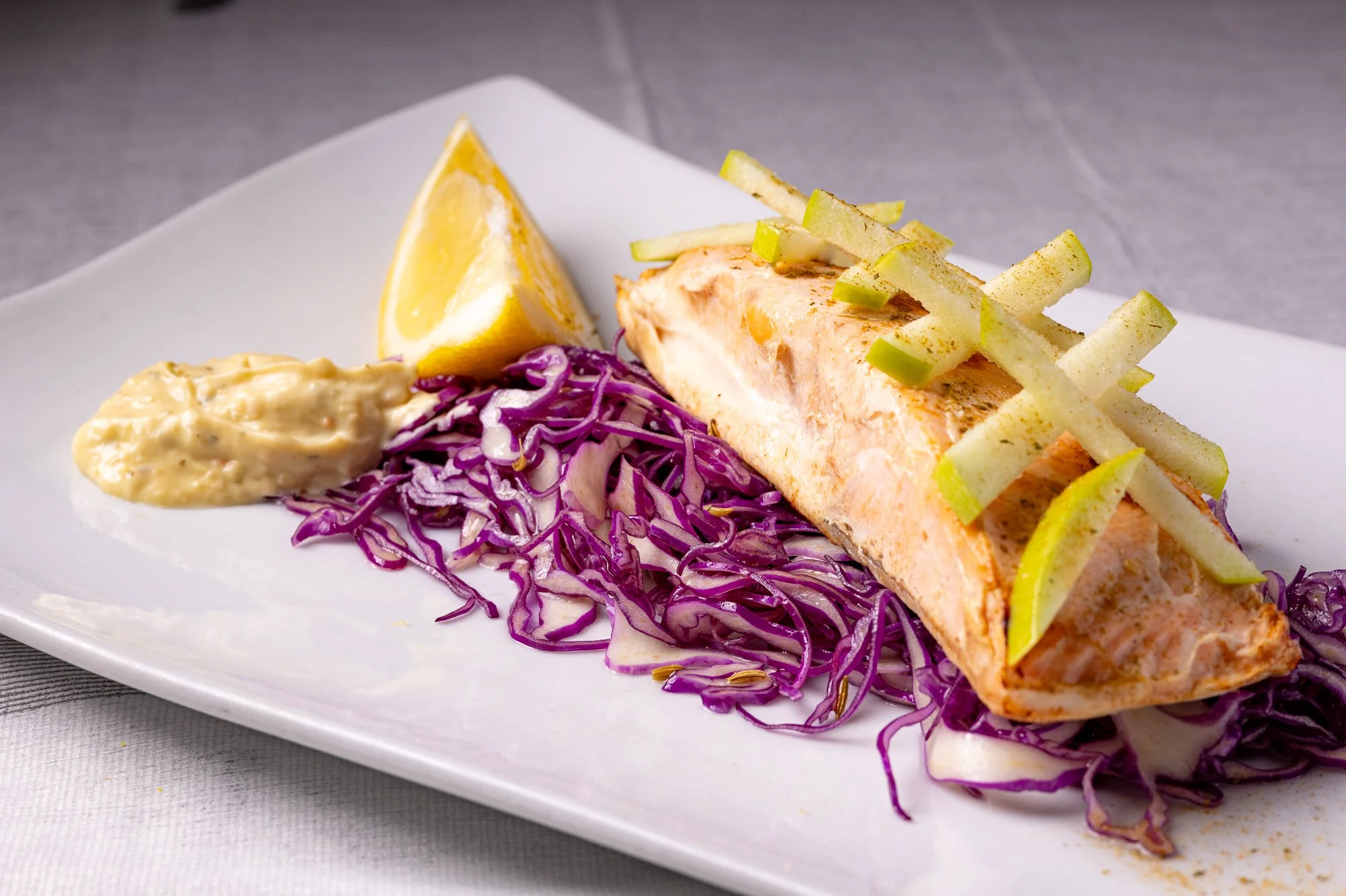 Filetto di salmone con fette di mela, accompagnato da insalata di cavolo viola e una fetta di limone, servito su un piatto bianco.