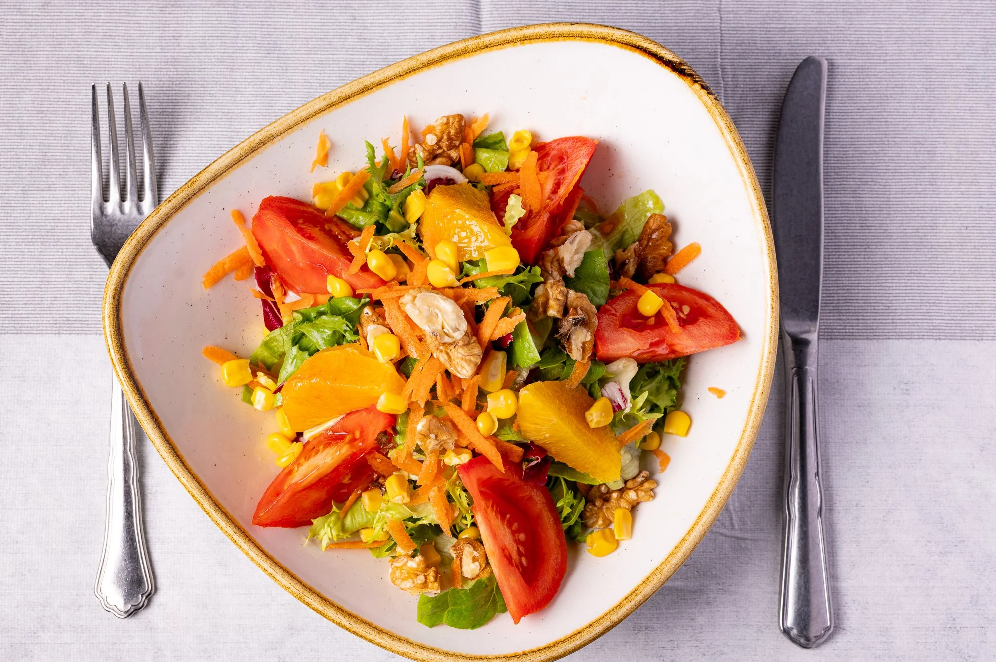 Insalata mista con lattuga, pomodori, carote, mais, arance e noci, servita in un piatto bianco con bordo marrone, accompagnata da una forchetta e un coltello di fronte, su un tovagliolo grigio chiaro.