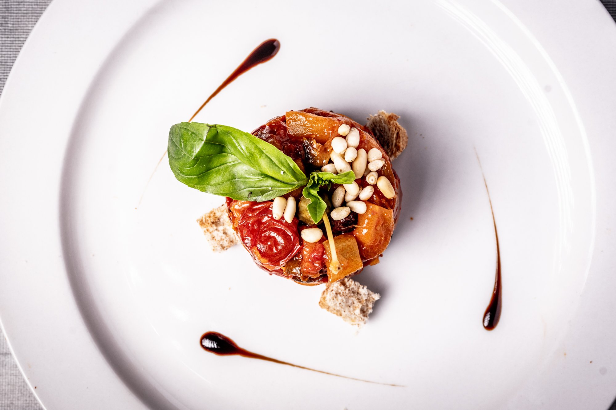 Piatto di caponata di mele con pomodoro, basilico fresco, pinoli, aceto balsamico e pane croccante, decorato con salsa di aceto balsamico.
