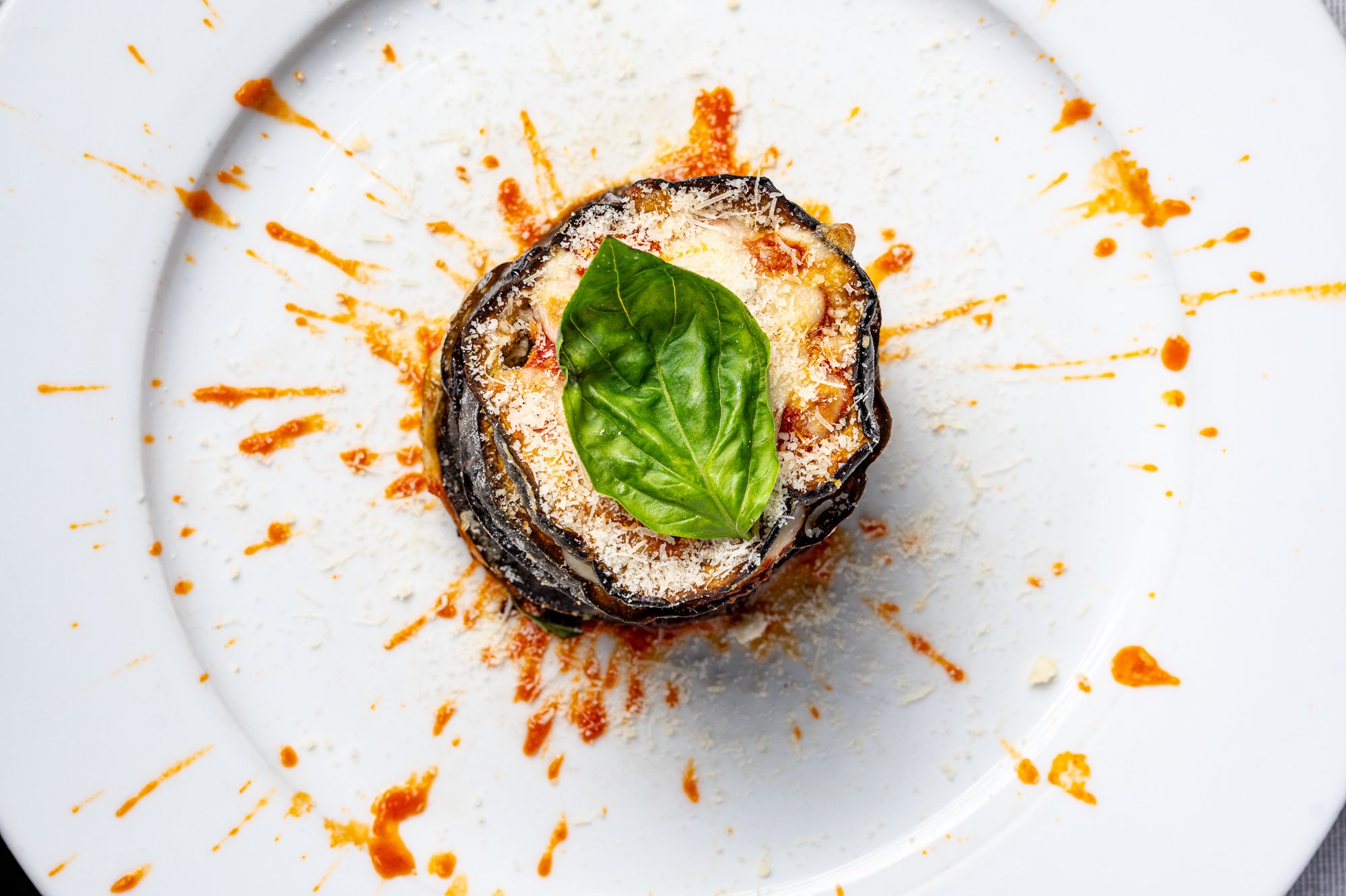Parmigiana di melanzane, decorata con foglia di basilico e parmigiano, servita su un piatto bianco con salsa di pomodoro.