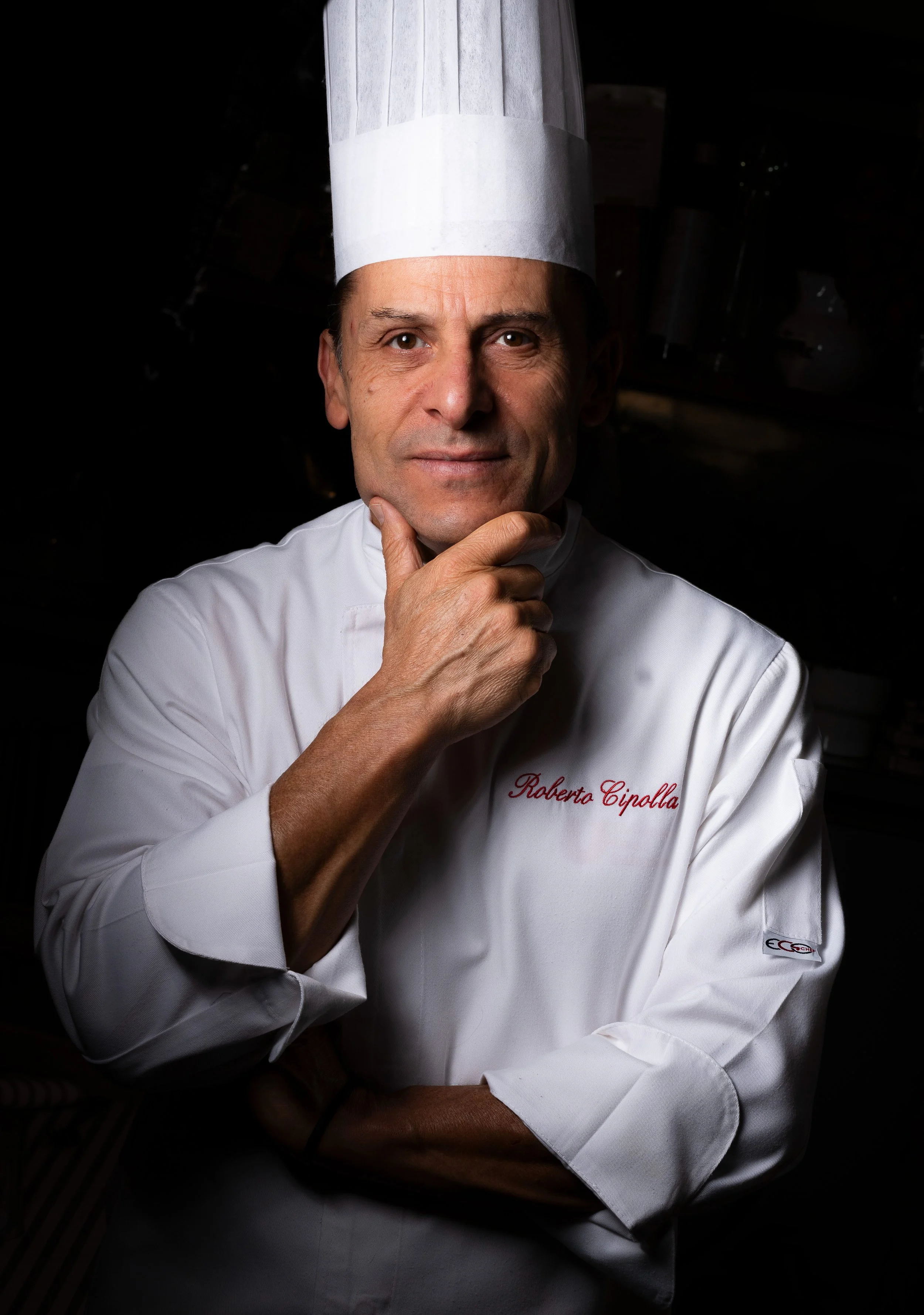 Chef Roberto Cipolla con toque bianco e giacca bianca, pensieroso, con mano sul mento, sfondo scuro.