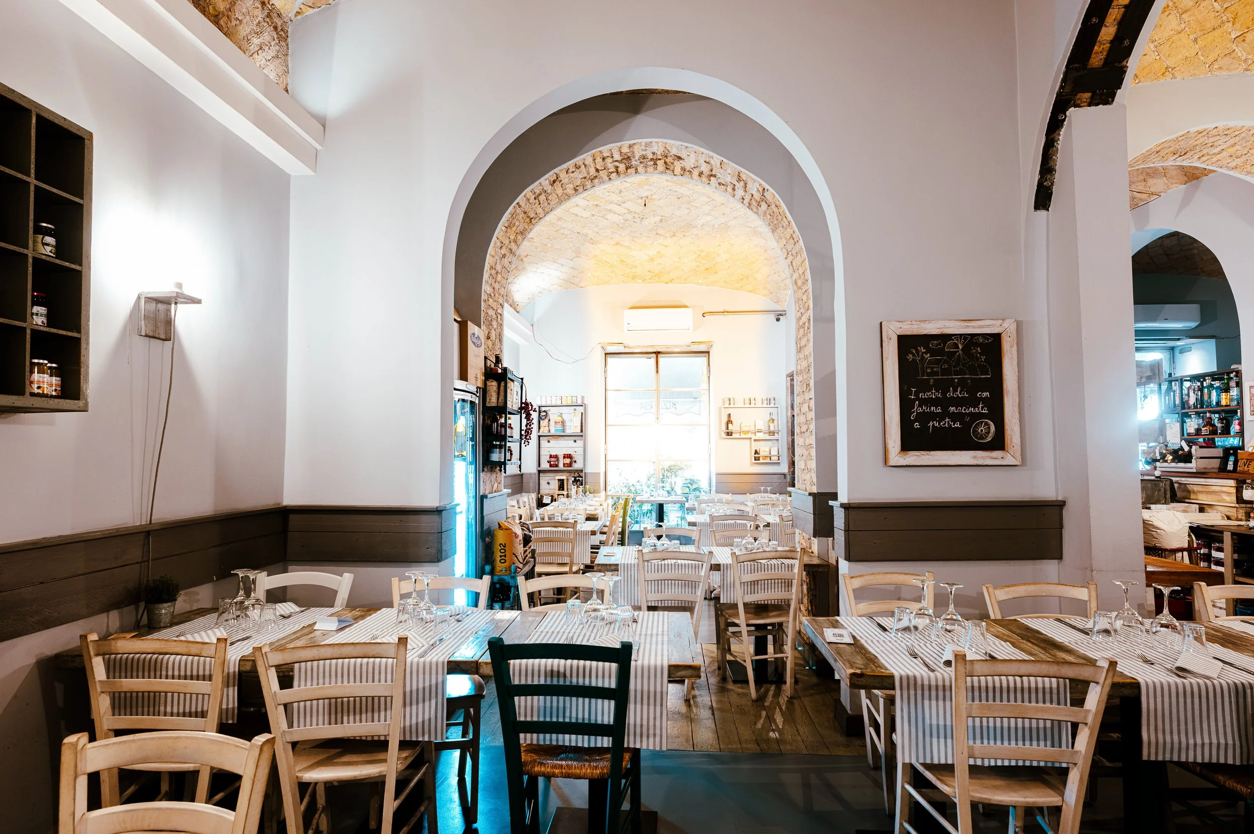 Interno di un ristorante con tavoli apparecchiati, sedie di legno e pareti bianche con archi in pietra.
