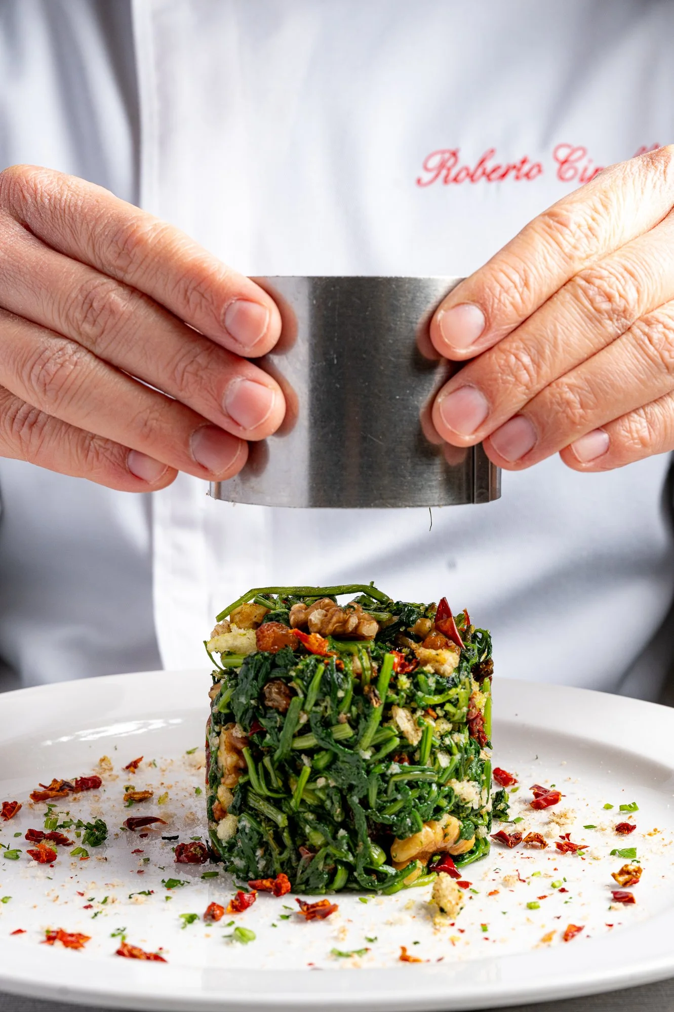Chef che utilizza uno stampo per formare un piatto di cicoria ripassata, decorato con spezie e peperoncino tritato.
