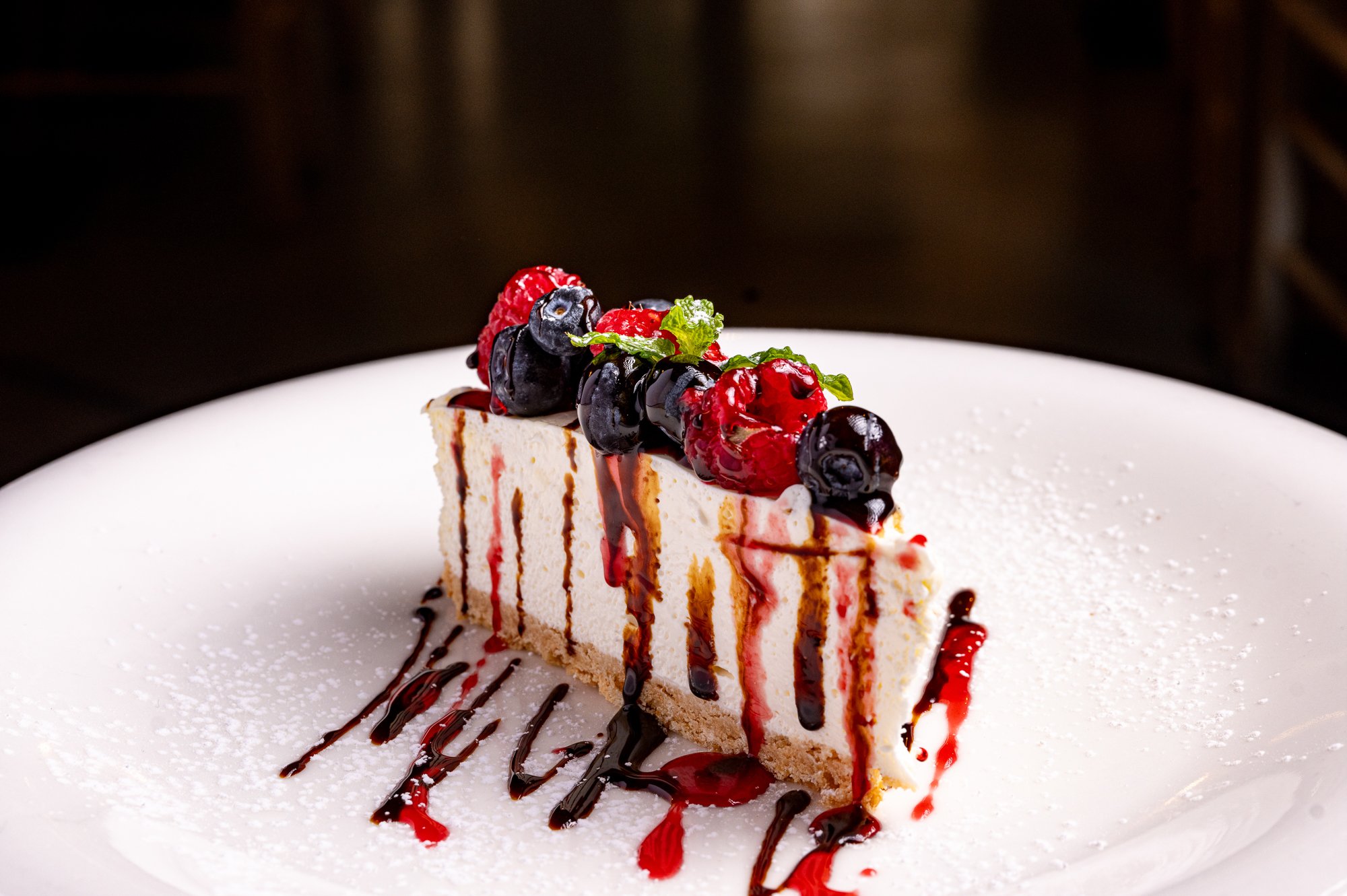 Torta cheesecake decorata con mirtilli, lamponi e foglie di menta, con salsa di cioccolato e lampone, servita su un piatto bianco con una leggera spolverata di zucchero a velo.