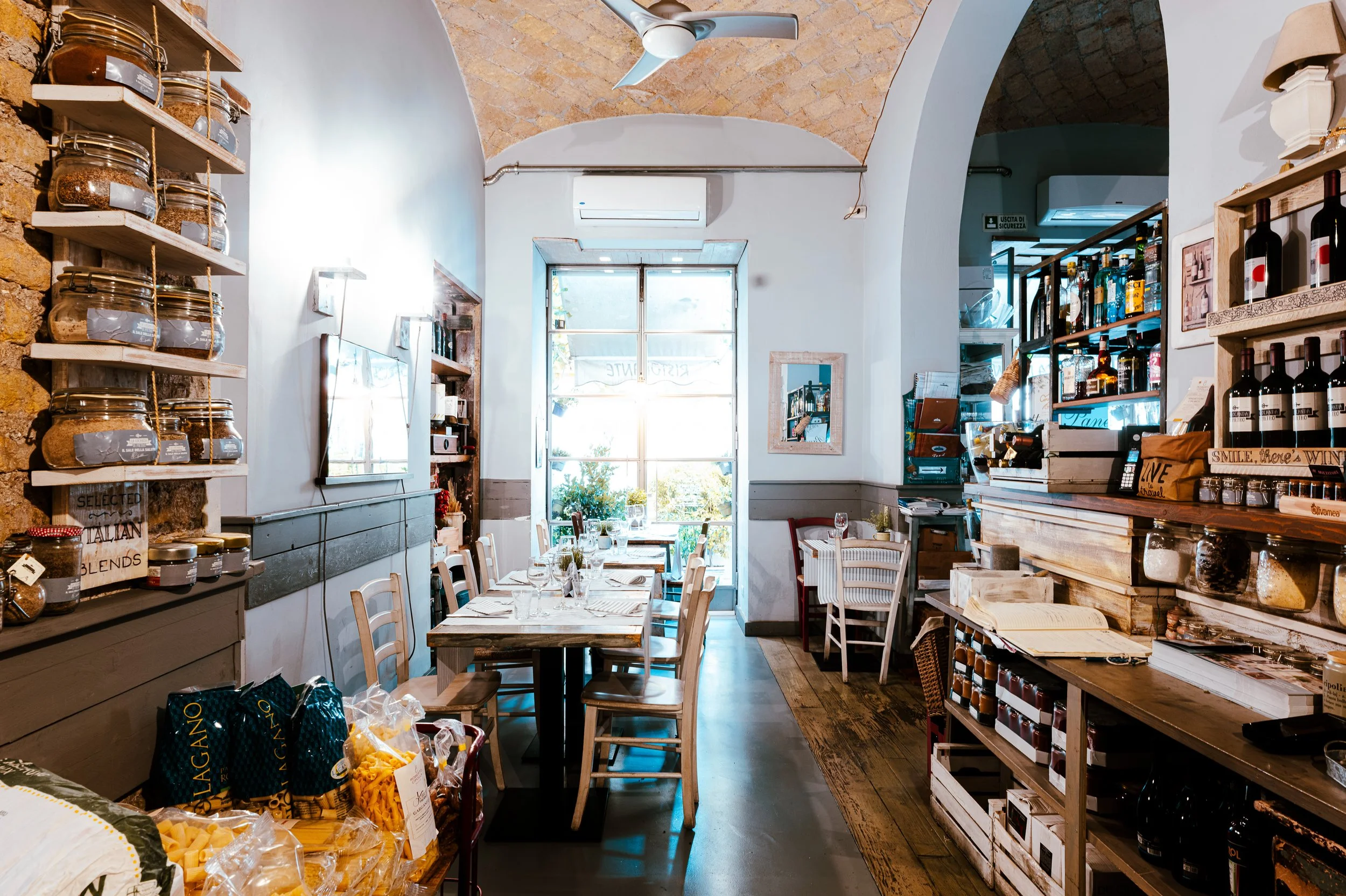 Interno di un ristorante con tavoli apparecchiati, scaffali con prodotti alimentari e bar con bottiglie e prodotti alimentari.