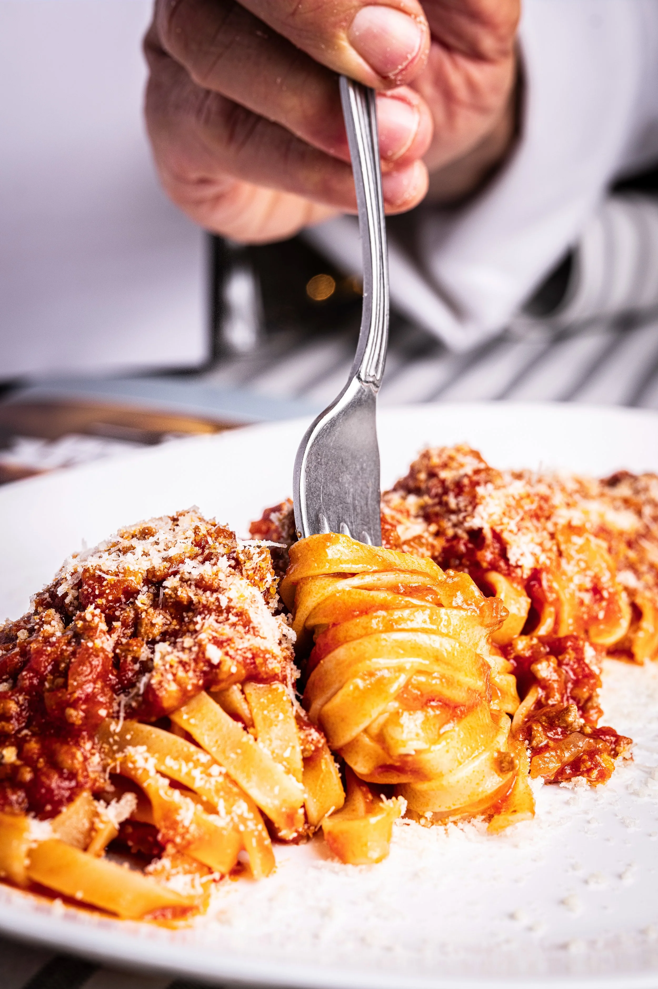 Mano che arrotola un piatto di pasta con ragù e Parmigiano Reggiano grattugiato su un piatto bianco.