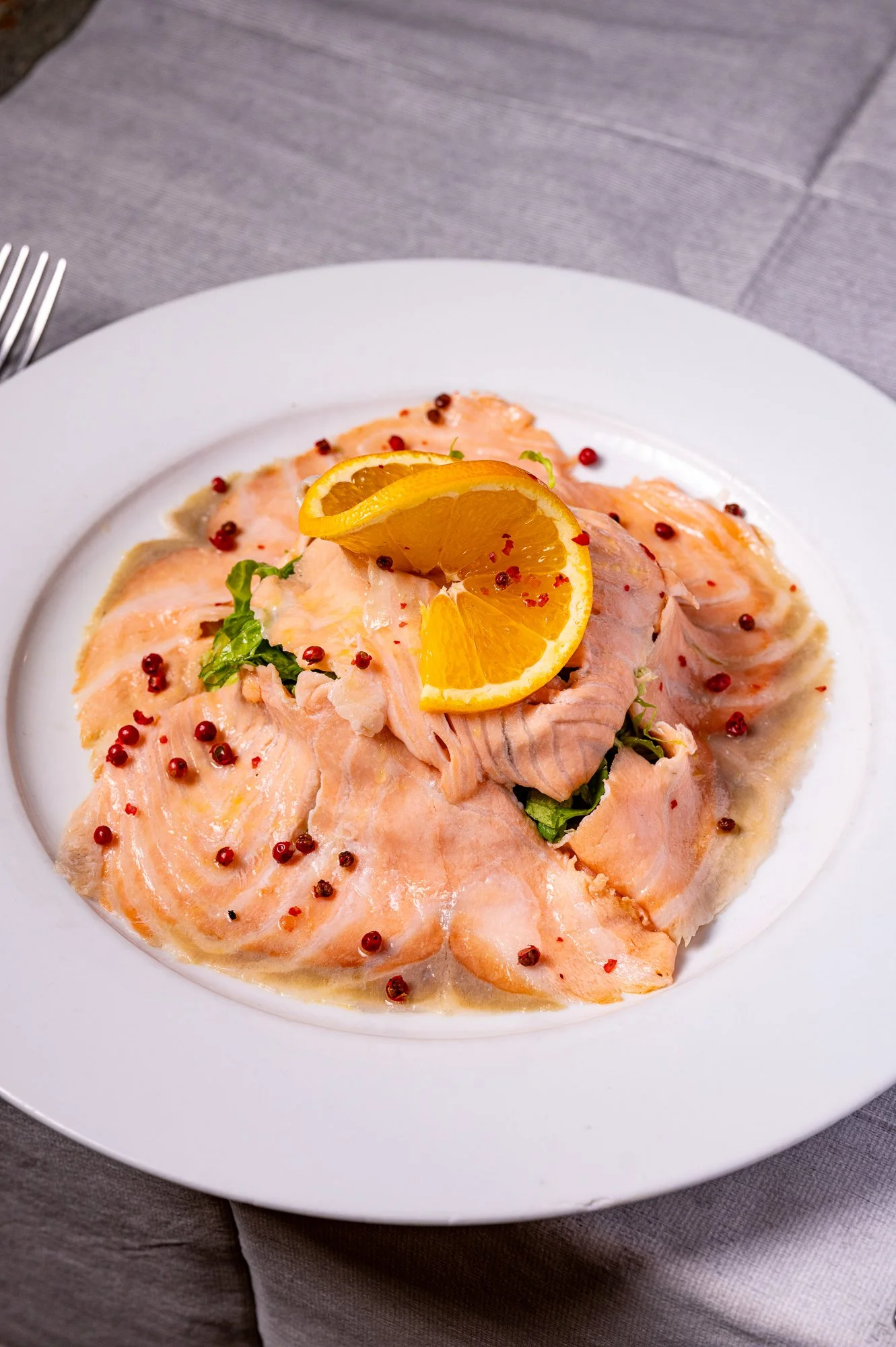 Salmone marinato agli agrumi, decorato con fette di arancia e pepe rosa, servito su un piatto bianco.