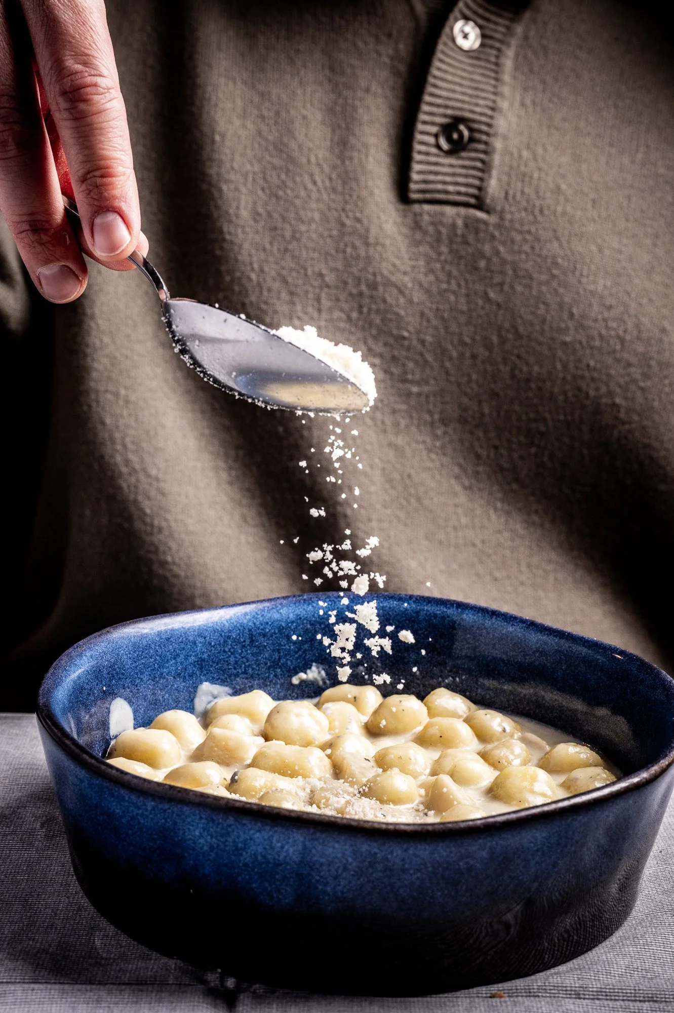 Una mano che sparge del Parmigiano Reggiano sopra un piatto di gnocchi al gorgonzola serviti in una ciotola blu.