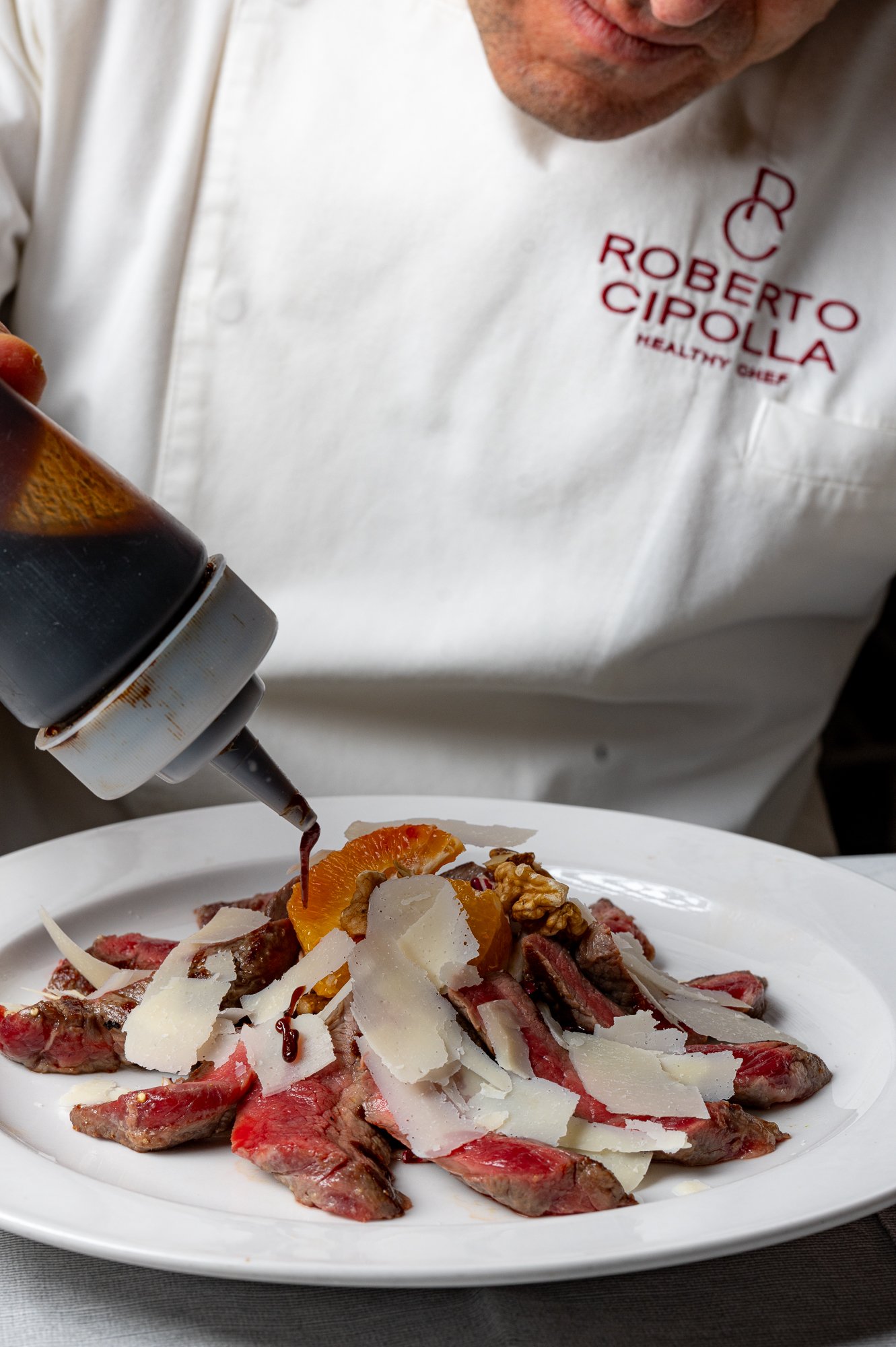Chef con camicia bianca con logo e nome, versa salsa su un piatto con tagliata di carne e condimenti.