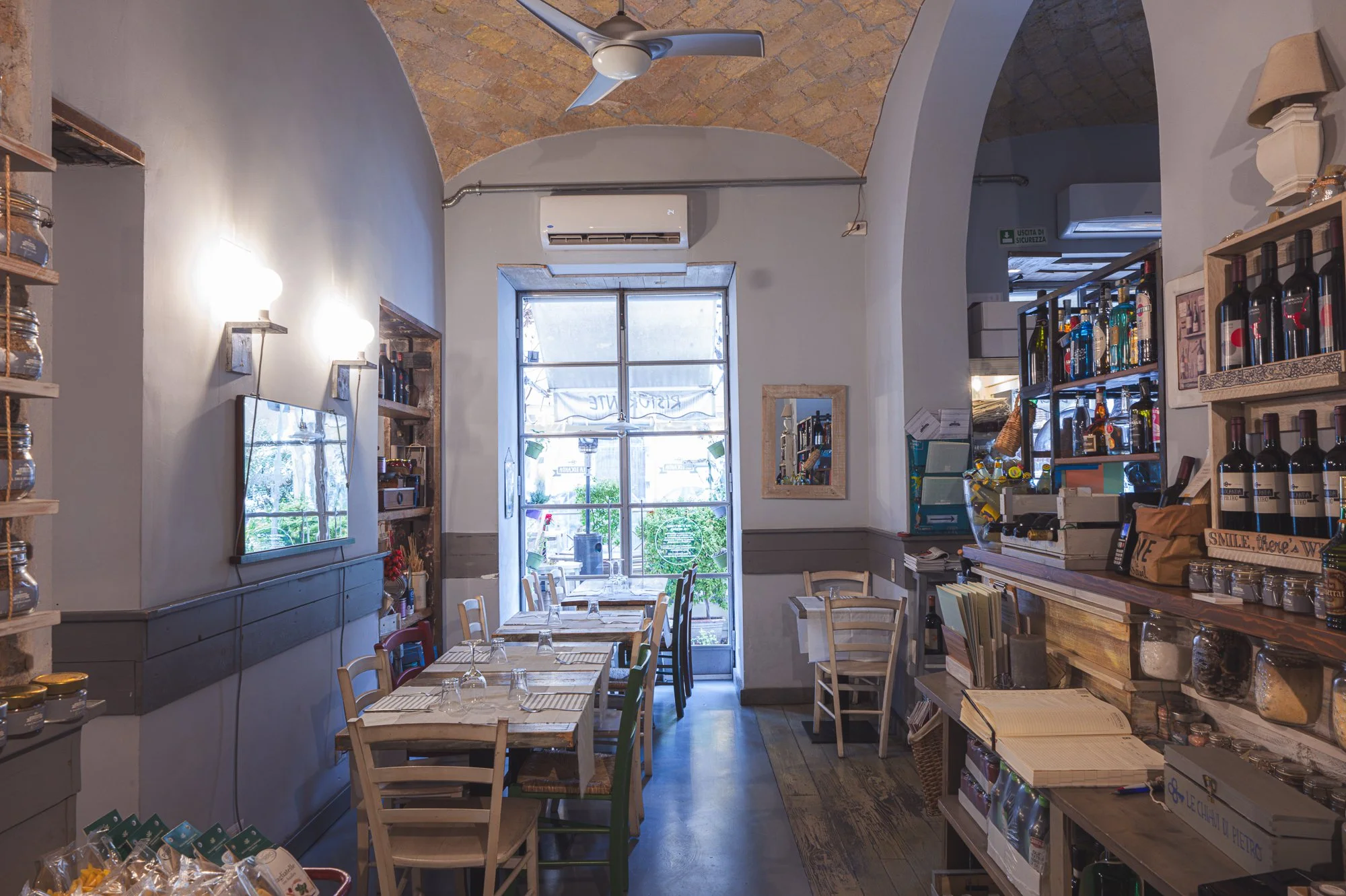 Interno di un ristorante con tavoli in legno e sedie, scaffali con bottiglie di vino e altri prodotti, e una grande finestra che lascia entrare luce naturale.