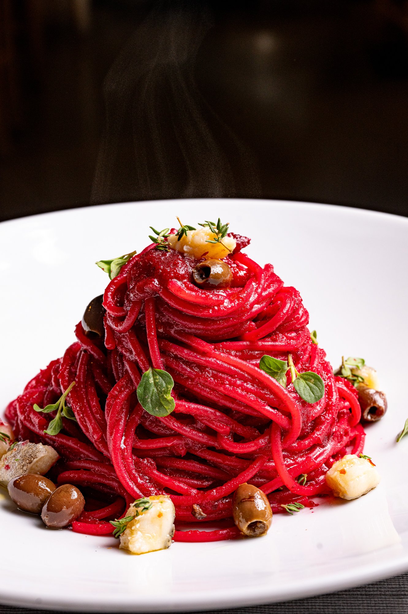 Spaghetti di barbabietola rossa con baccalà. Presentati su un piatto bianco, sfondo nero.