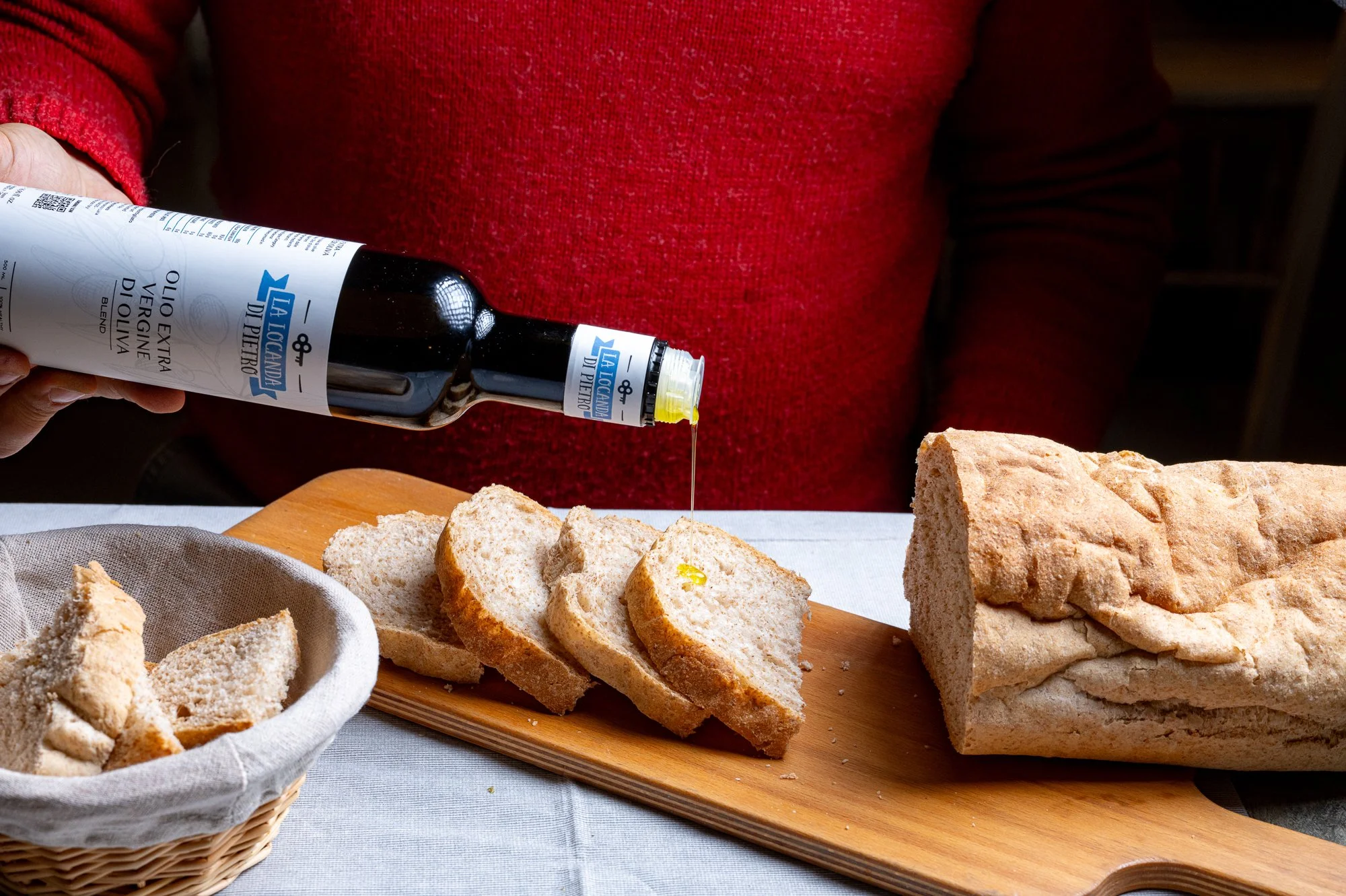 Una bottiglia di olio extravergine di oliva di marca 'La Locanda di Pietro' viene versata su fette di pane bianco su un tagliere di legno, con pezzi di pane in una ciotola di vimini accanto.